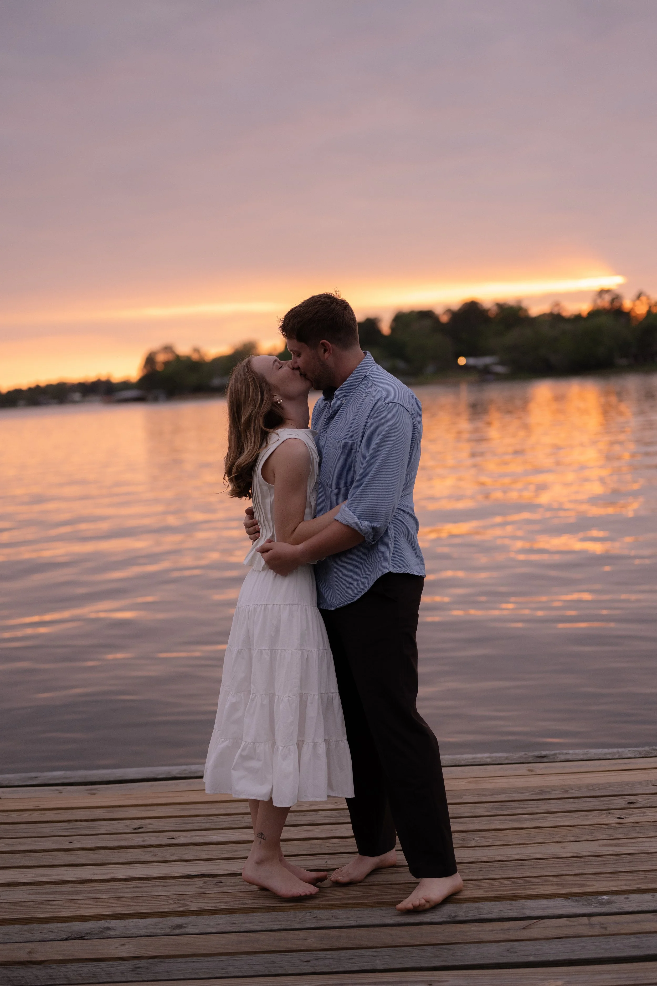 A couple kissing on a wooden dock by a lake during sunset, with trees and a colorful sky in the background.