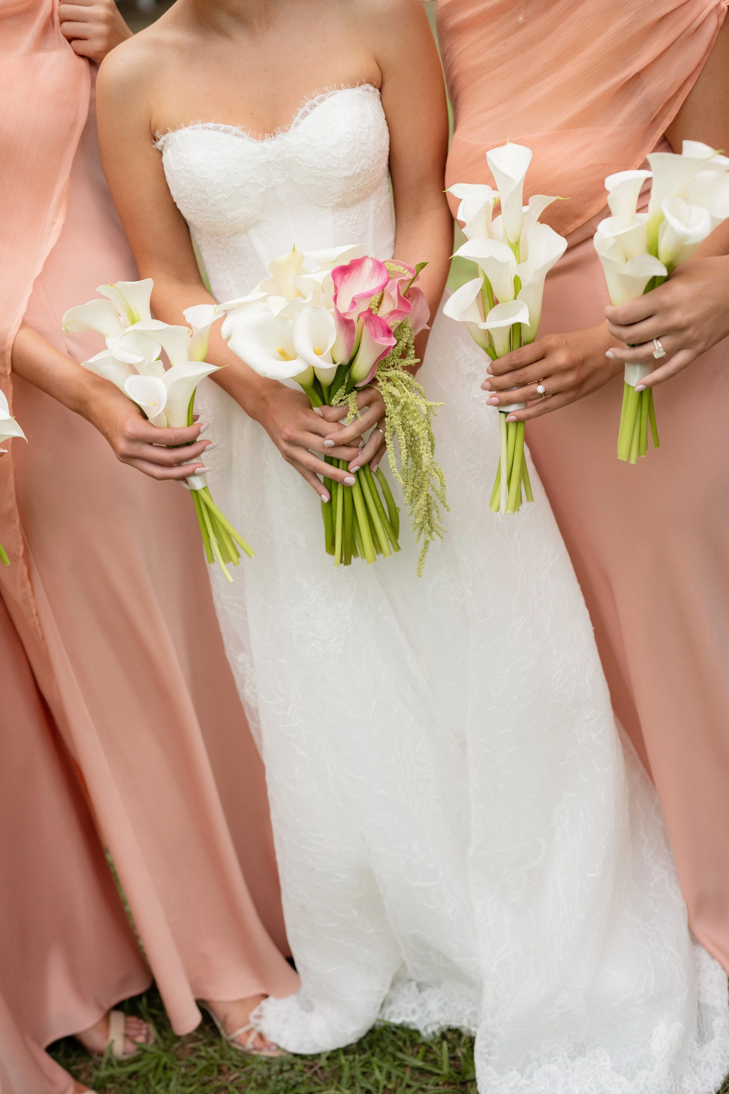 Close-up of women holding bouquets of white calla lilies and pink flowers, standing on grass, with the focus on their dresses and hands.