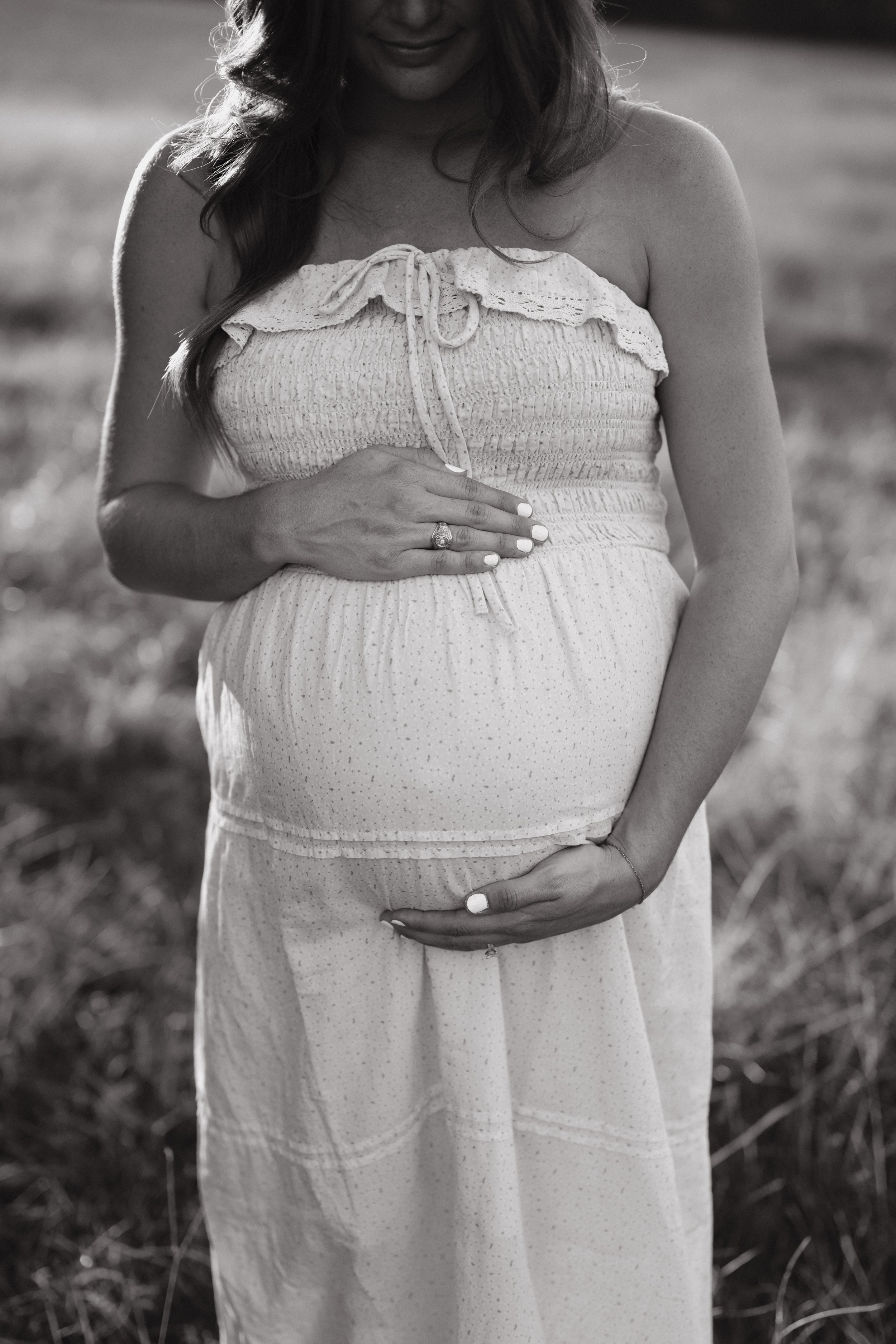 A pregnant woman in a strapless, light-colored dress with a ruffled top, holding her belly with one hand and resting the other on her stomach in an outdoor setting. The photo is in black and white.