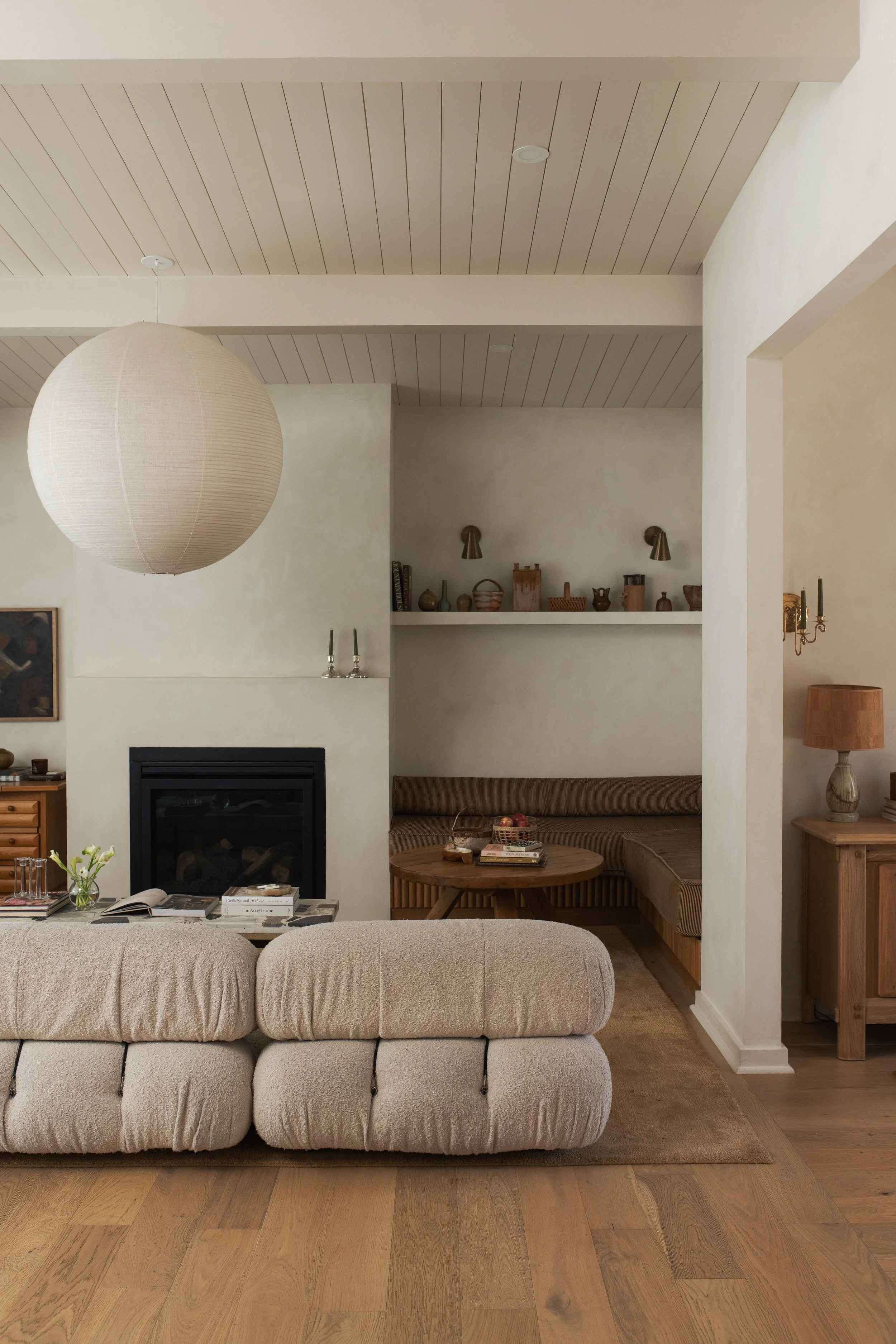 Living room with a beige upholstered sectional sofa, a wooden coffee table with books and a basket, a fireplace, a side cabinet with flowers, wall-mounted brass lamps, a floating shelf with pottery and books, a large paper lantern hanging from the ce