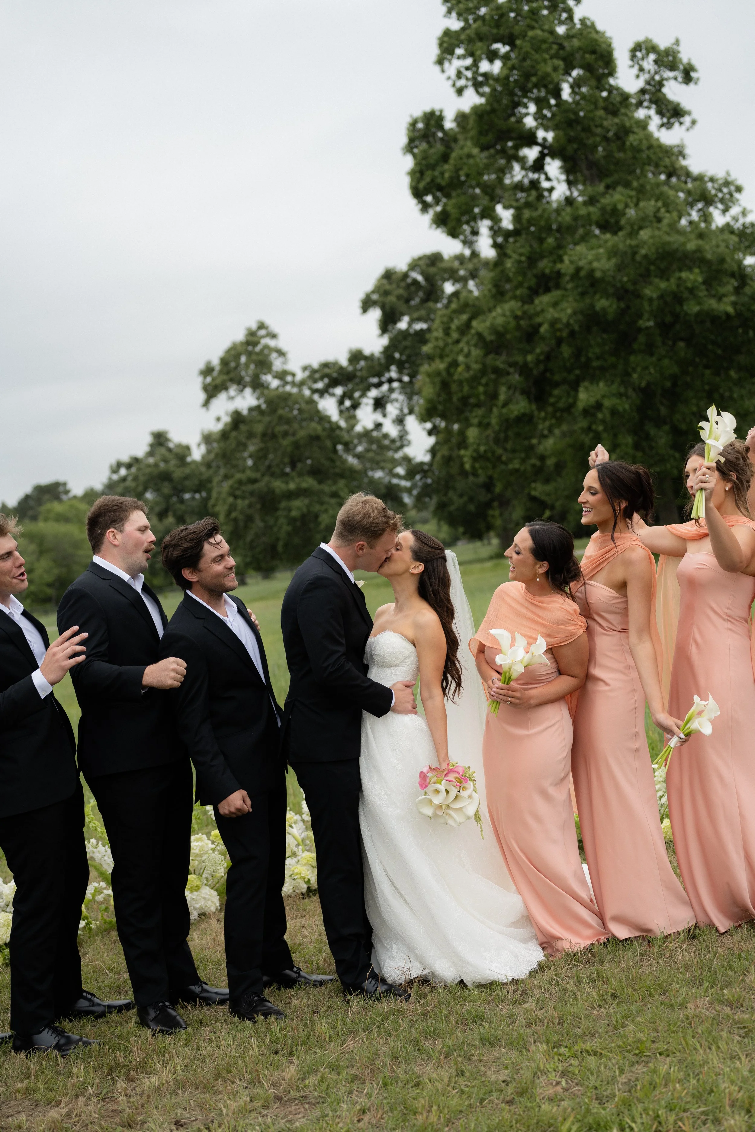 A wedding ceremony outdoors with the bride and groom kissing, surrounded by friends and bridesmaids holding flowers, on a grassy field with trees in the background.