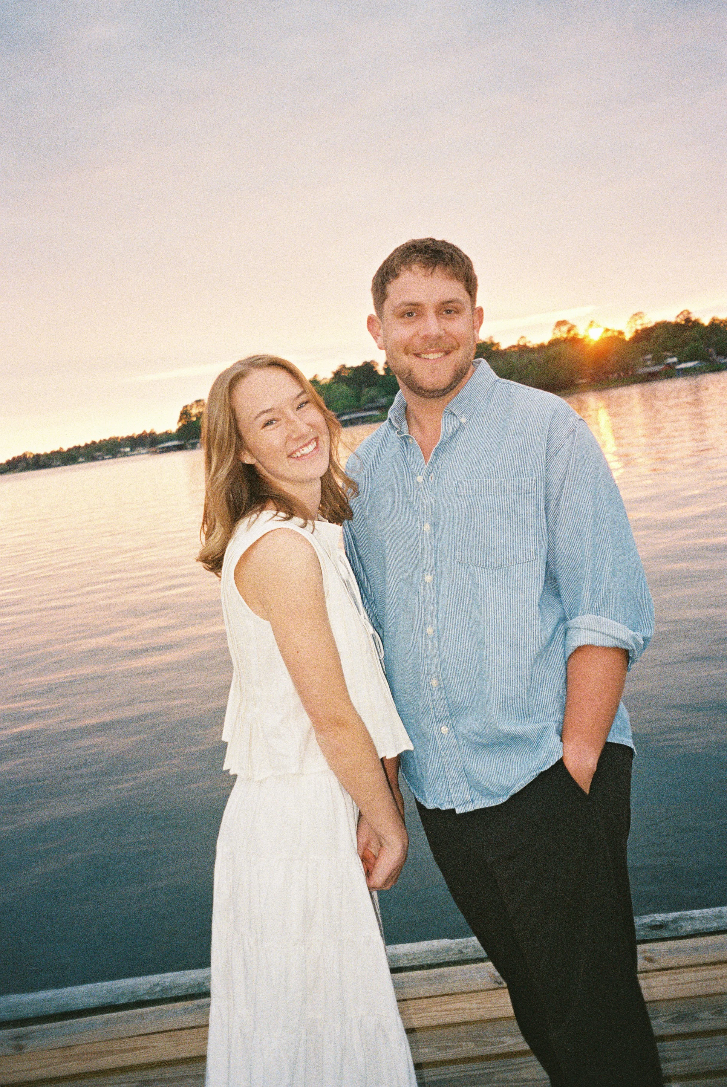 A smiling young woman and man stand hand in hand by a body of water during sunset, with trees and houses in the background.