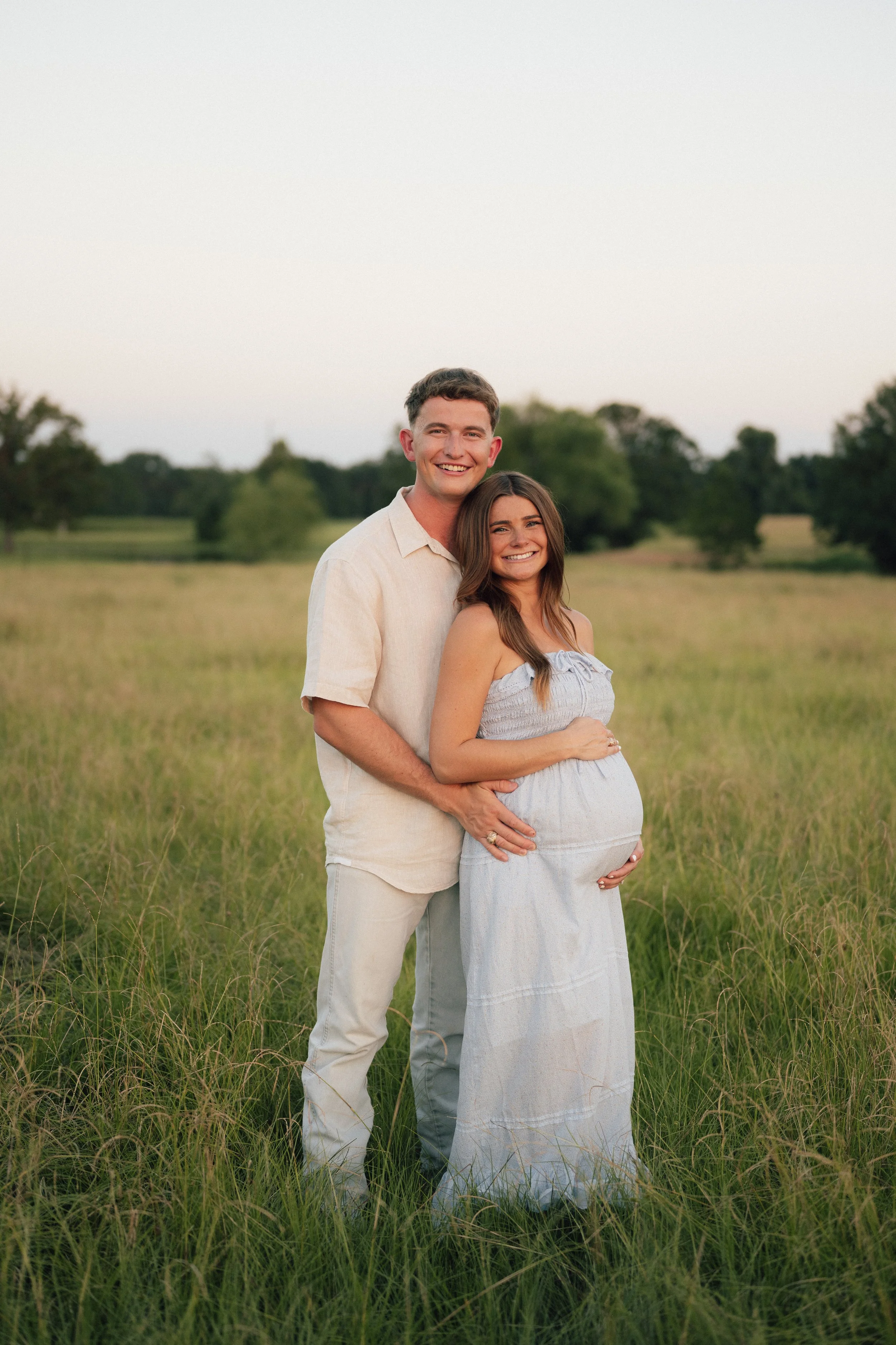 A couple standing in a grassy field, with the woman visibly pregnant, smiling at the camera, during sunset or late afternoon in a rural outdoor setting with trees in the background.