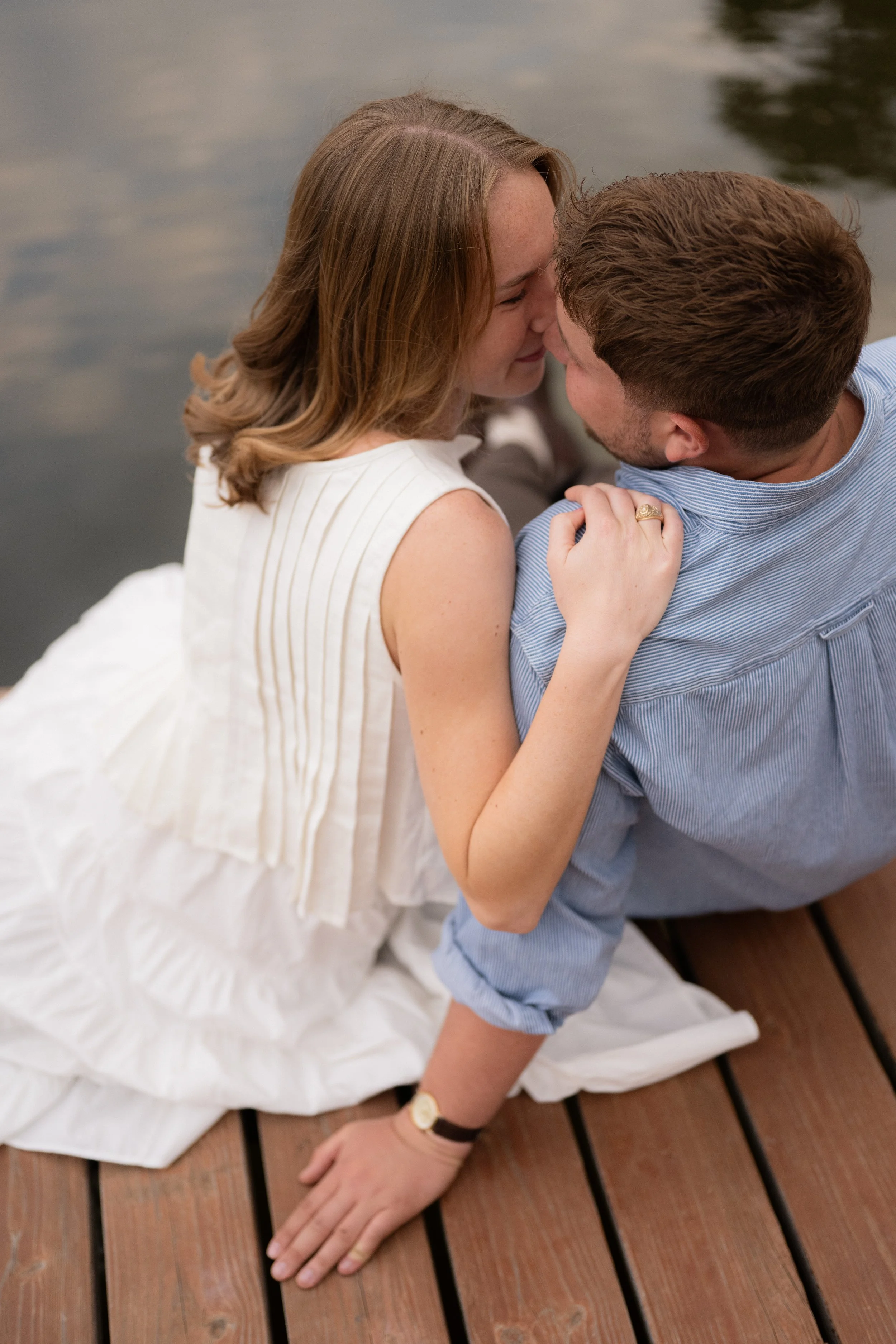 A woman and a man are sitting close together on a wooden dock, about to kiss. The woman has shoulder-length brown hair and is wearing a white dress, while the man has short brown hair and is wearing a blue striped shirt. The woman's left hand is rest