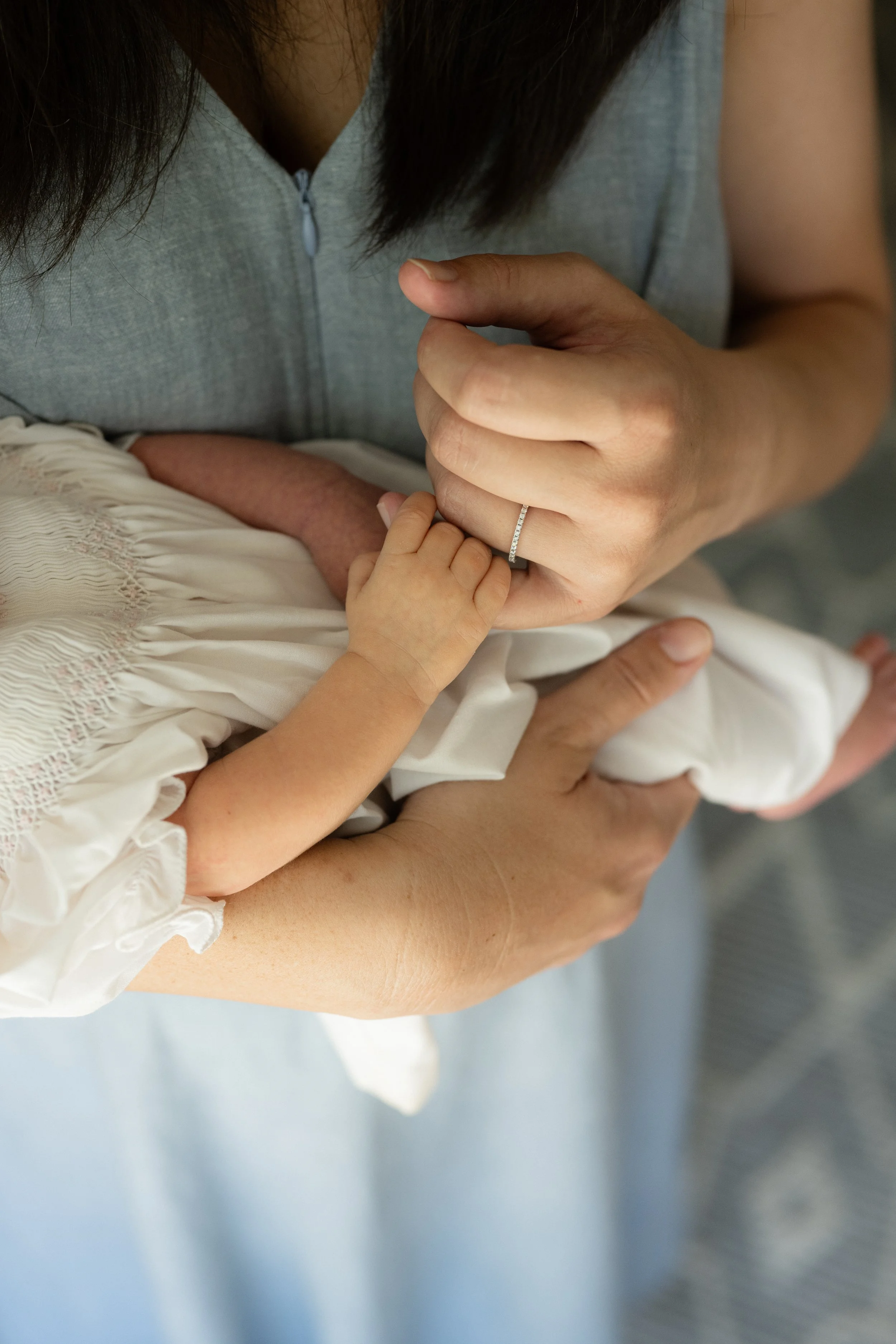 A woman holding a child's hand, showing a close-up of their hands together. The woman is wearing a ring and the child is wearing a white outfit with lace details.