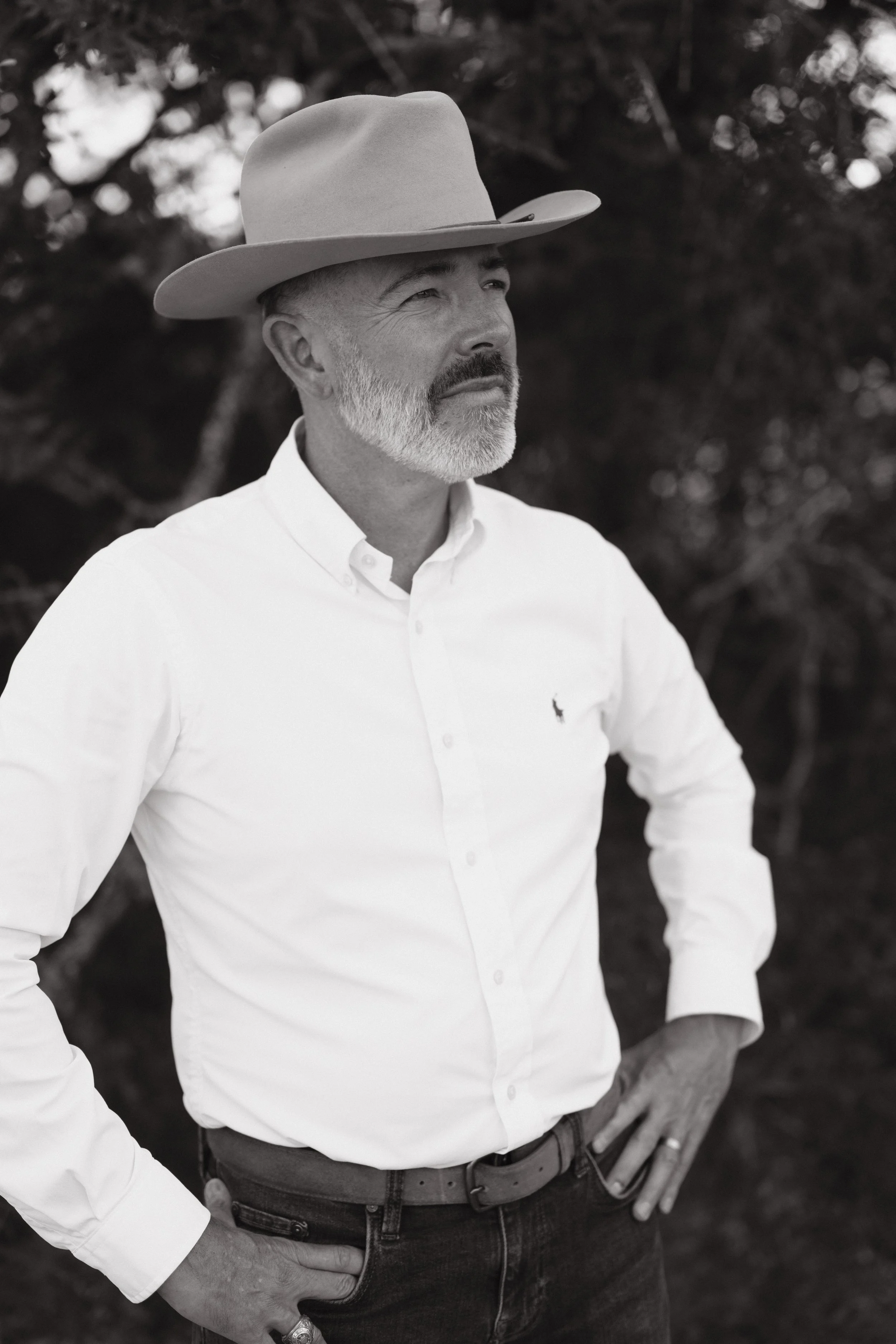 A black and white photo of a man with a beard and mustache wearing a wide-brimmed hat and a white button-down shirt. He is standing outdoors with trees in the background, with one hand resting on his hip.