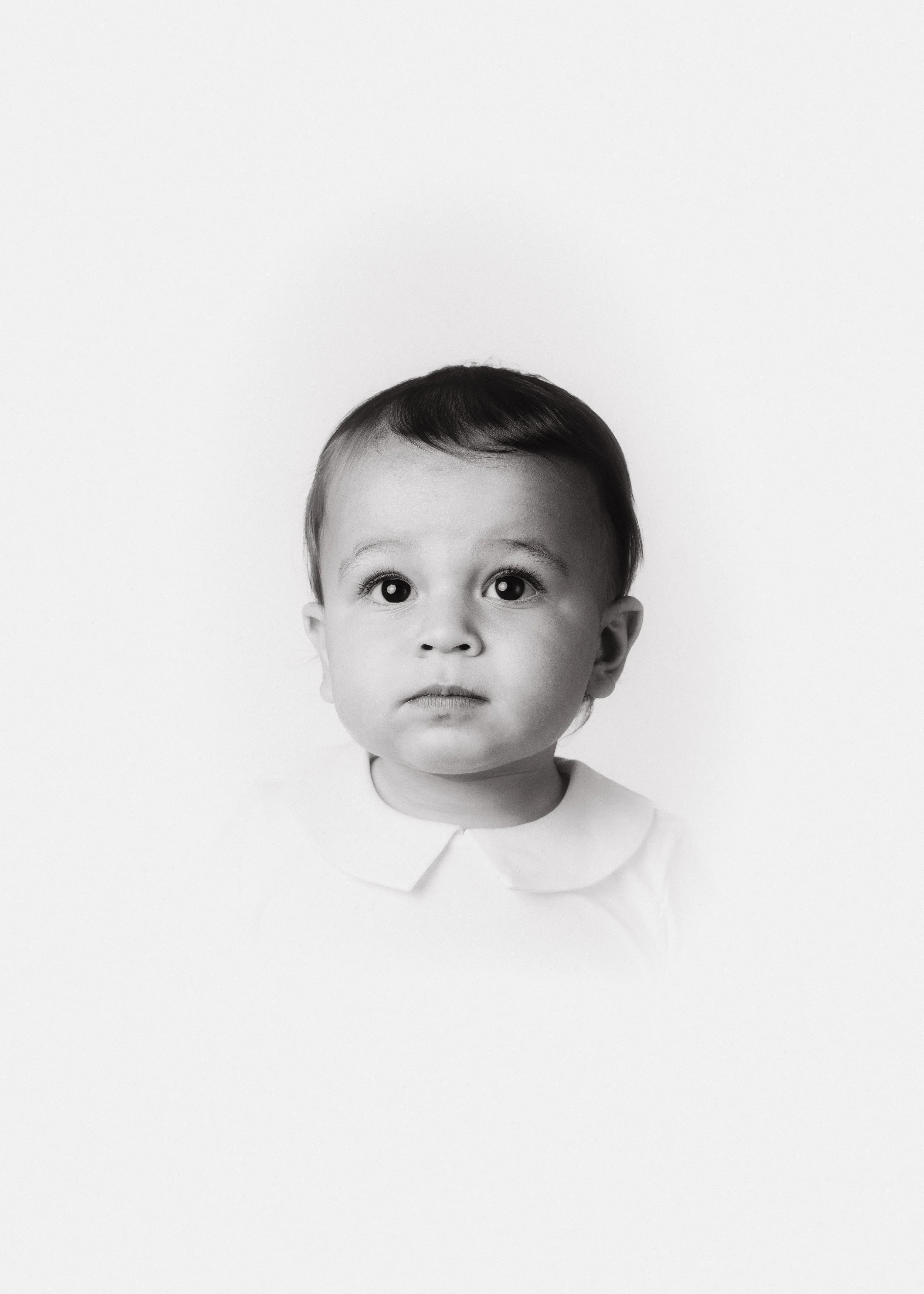 Black and white photo of a young child with big eyes, short dark hair, wearing a white shirt with a collar, against a plain light background.