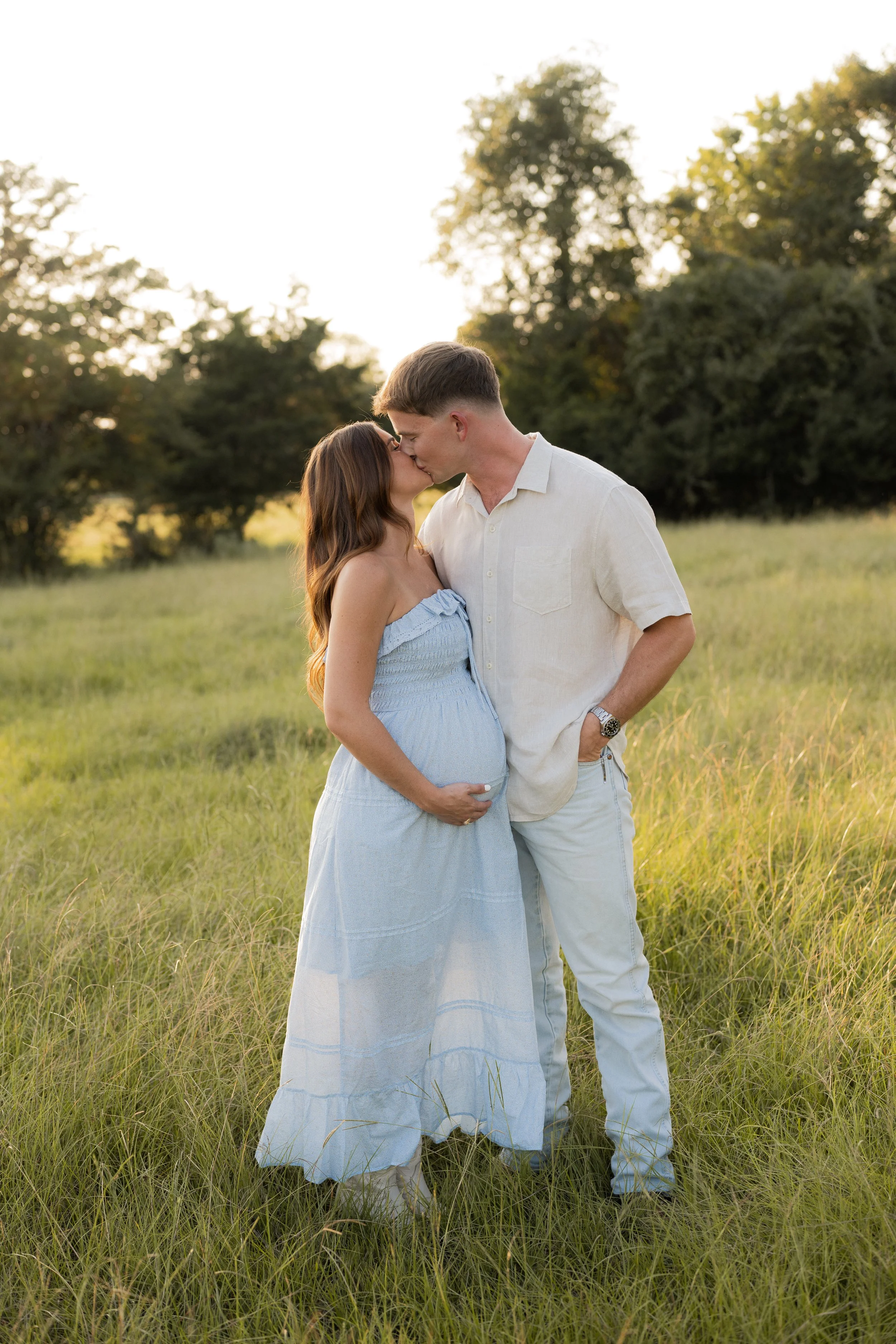 A couple, with the woman being pregnant, sharing a kiss in a field during sunset, surrounded by trees.