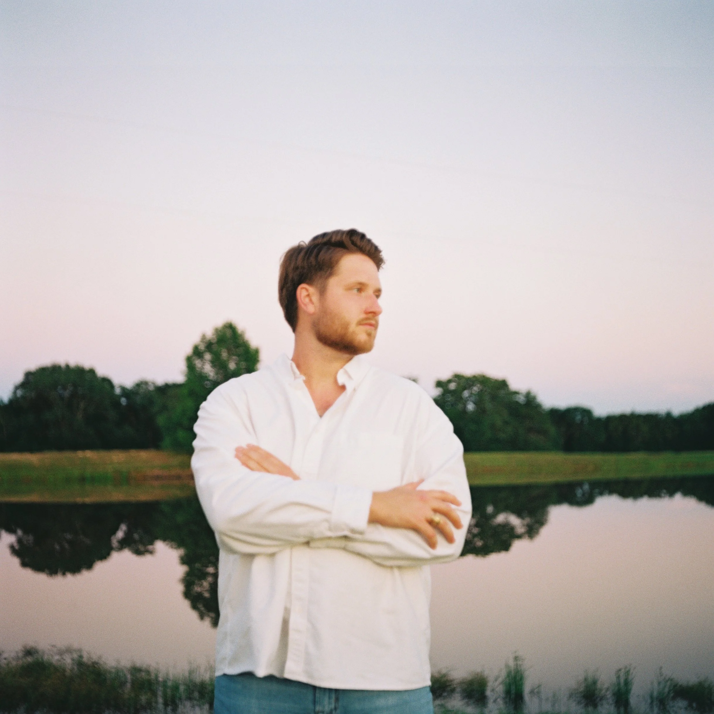 A man with brown hair and a beard standing outdoors near a water body and trees, wearing a white shirt and blue jeans, with arms crossed and looking to his right against a cloudy sky.