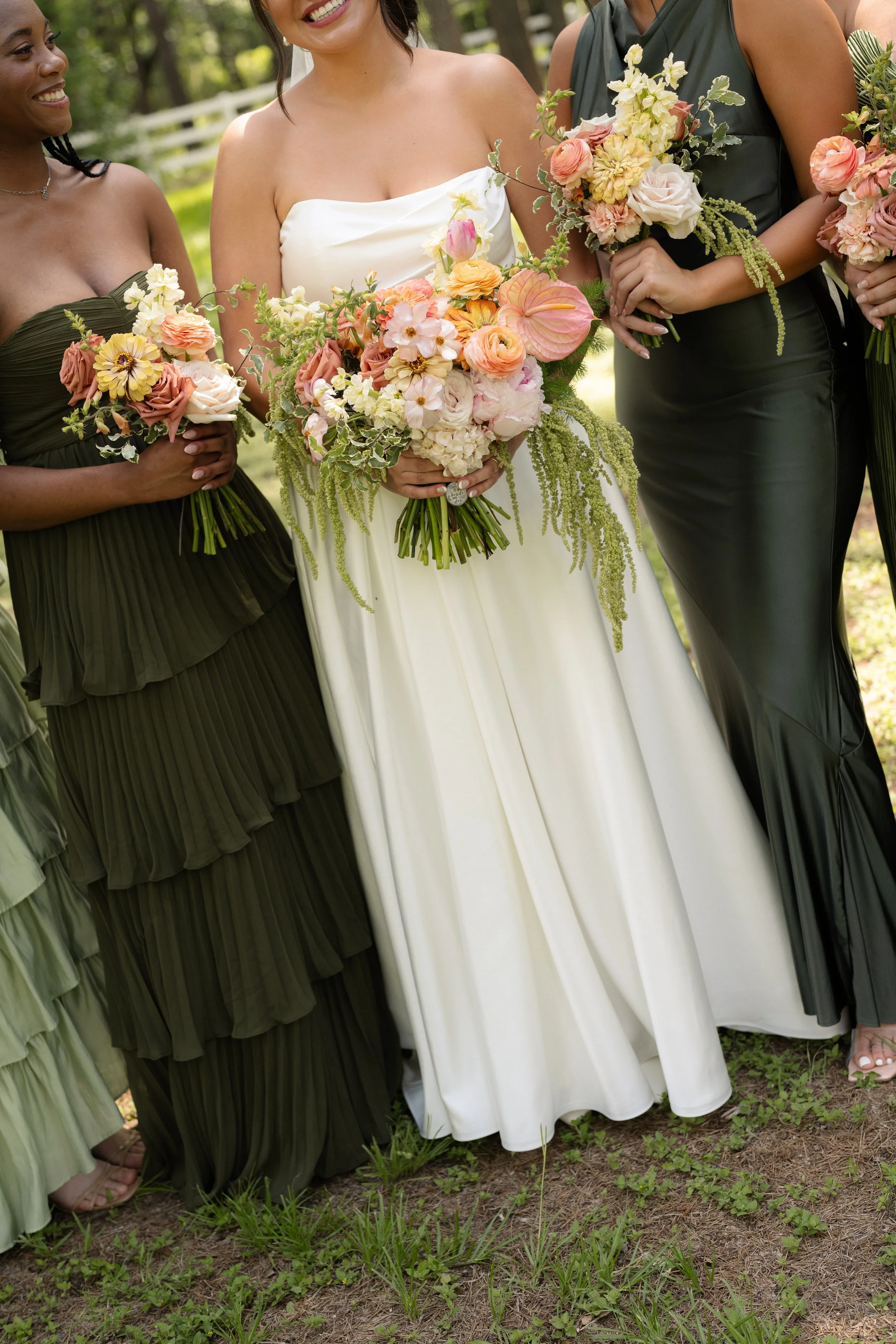 Close-up of three women standing outdoors, wearing formal dresses, holding bouquets of flowers, smiling, with green grass and trees in the background.
