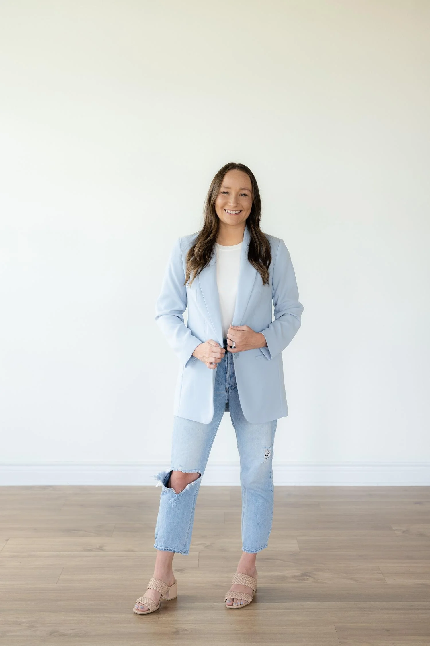 Woman with long, brown hair smiling, wearing a light blue blazer over a white shirt, ripped jeans, and beige high-heeled sandals, standing against a plain white wall with wooden flooring.