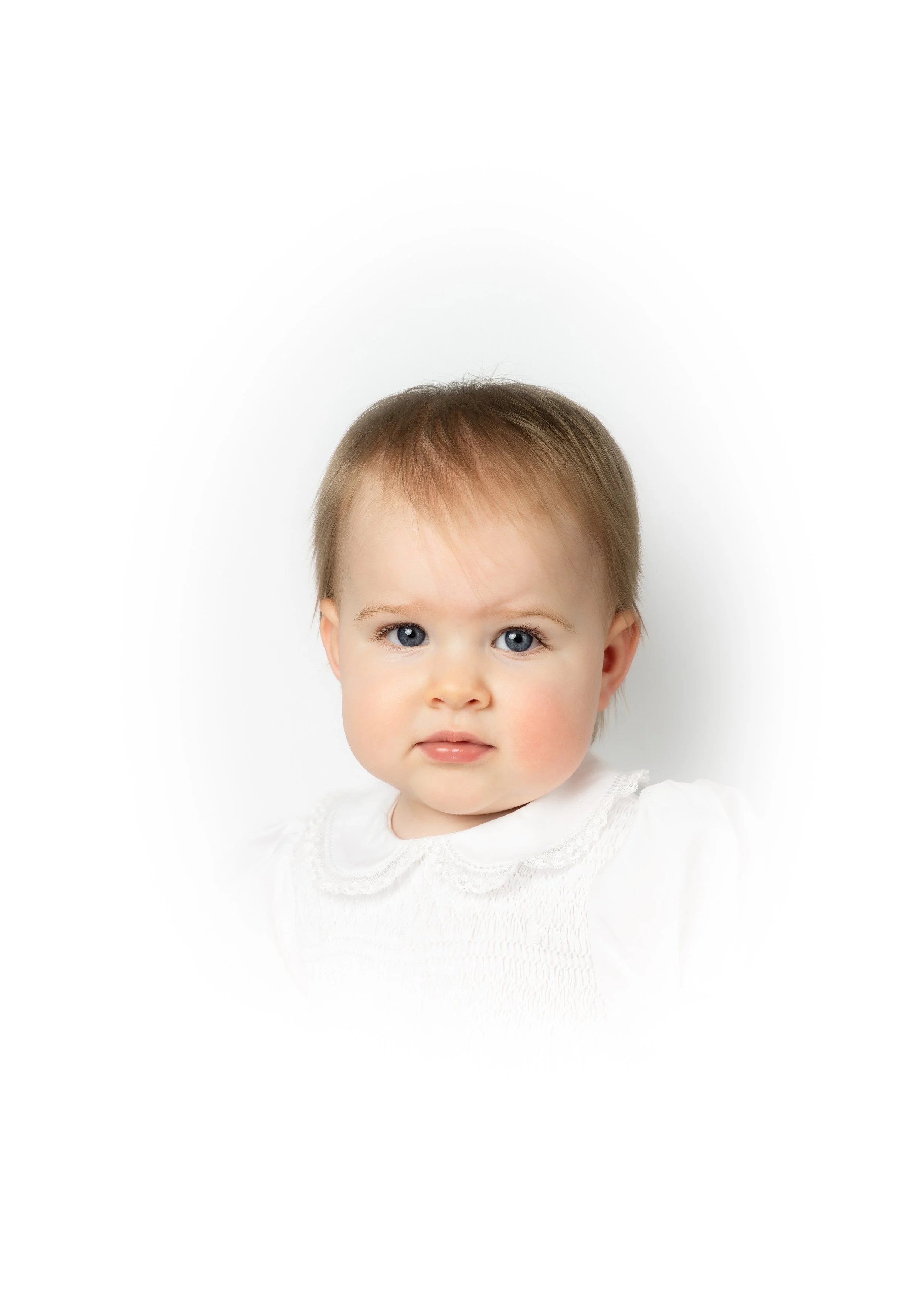 Close-up portrait of a young child with blue eyes, light brown hair, wearing a white top, against a plain white background.