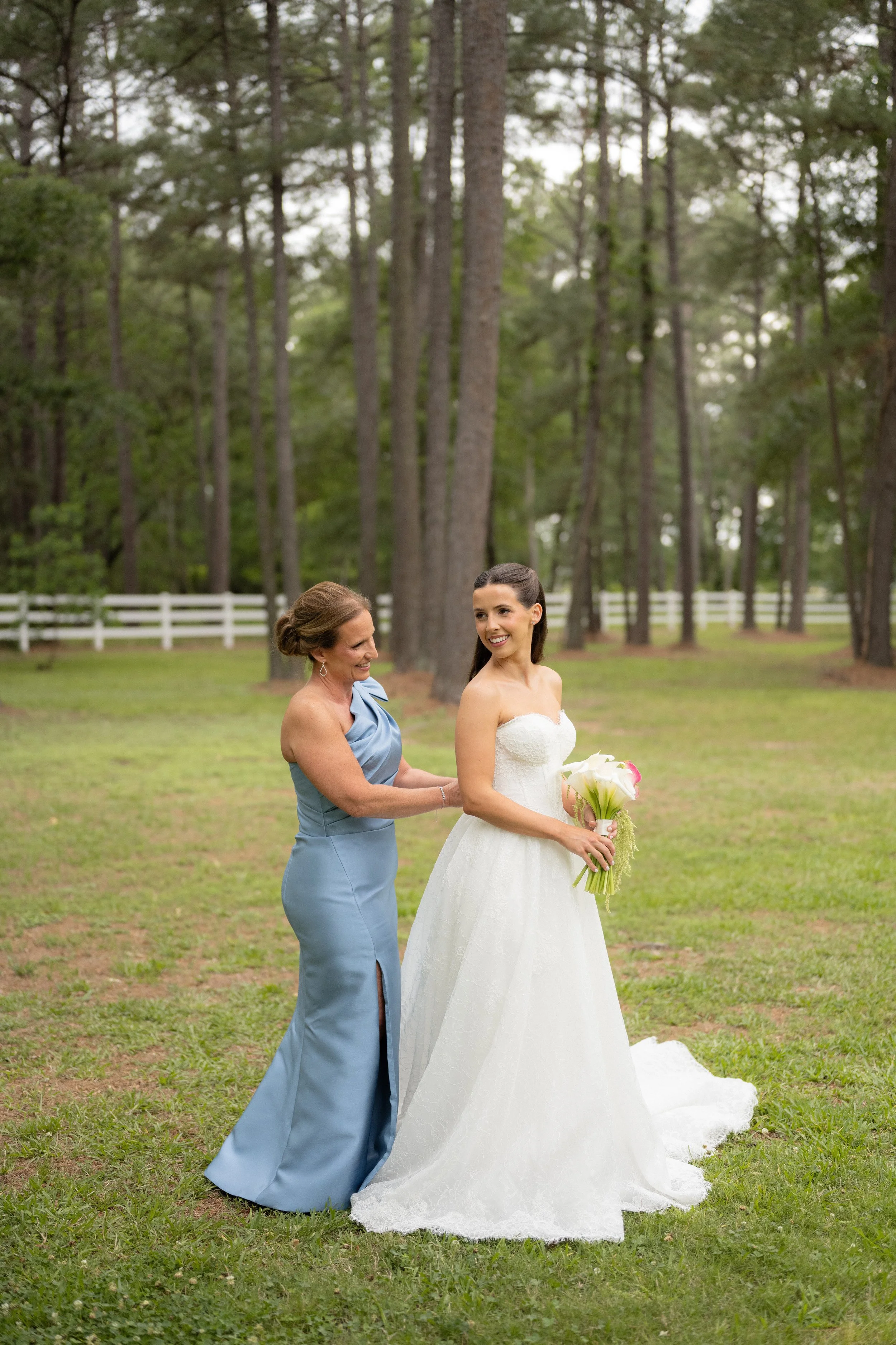 A bride in a white wedding dress standing outdoors on grass, smiling and holding a bouquet, with an older woman in a light blue dress assisting her, in a wooded area with tall trees and a white fence in the background.