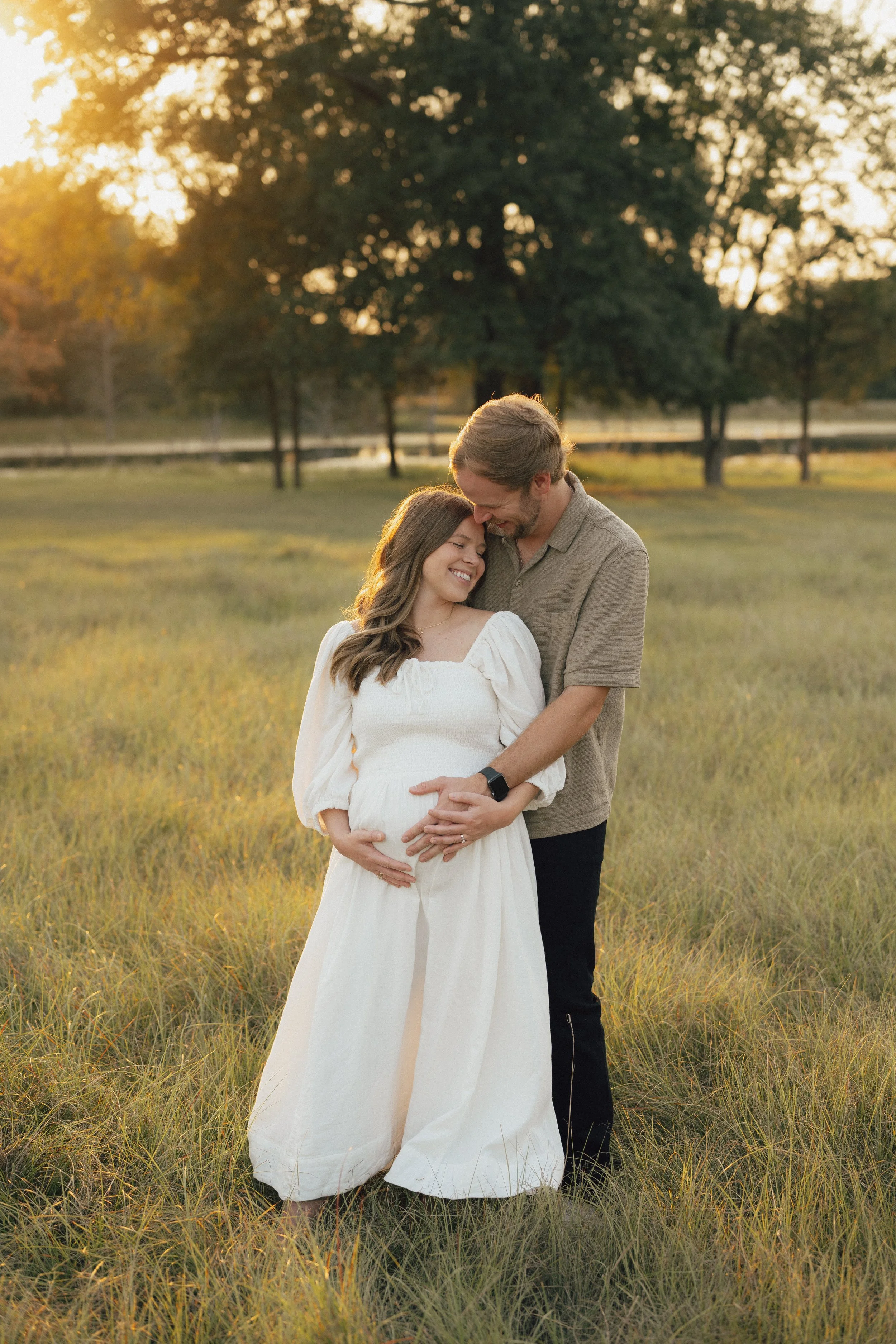 A man and woman standing in a grassy field during sunset, embracing each other with the woman visibly pregnant and smiling.