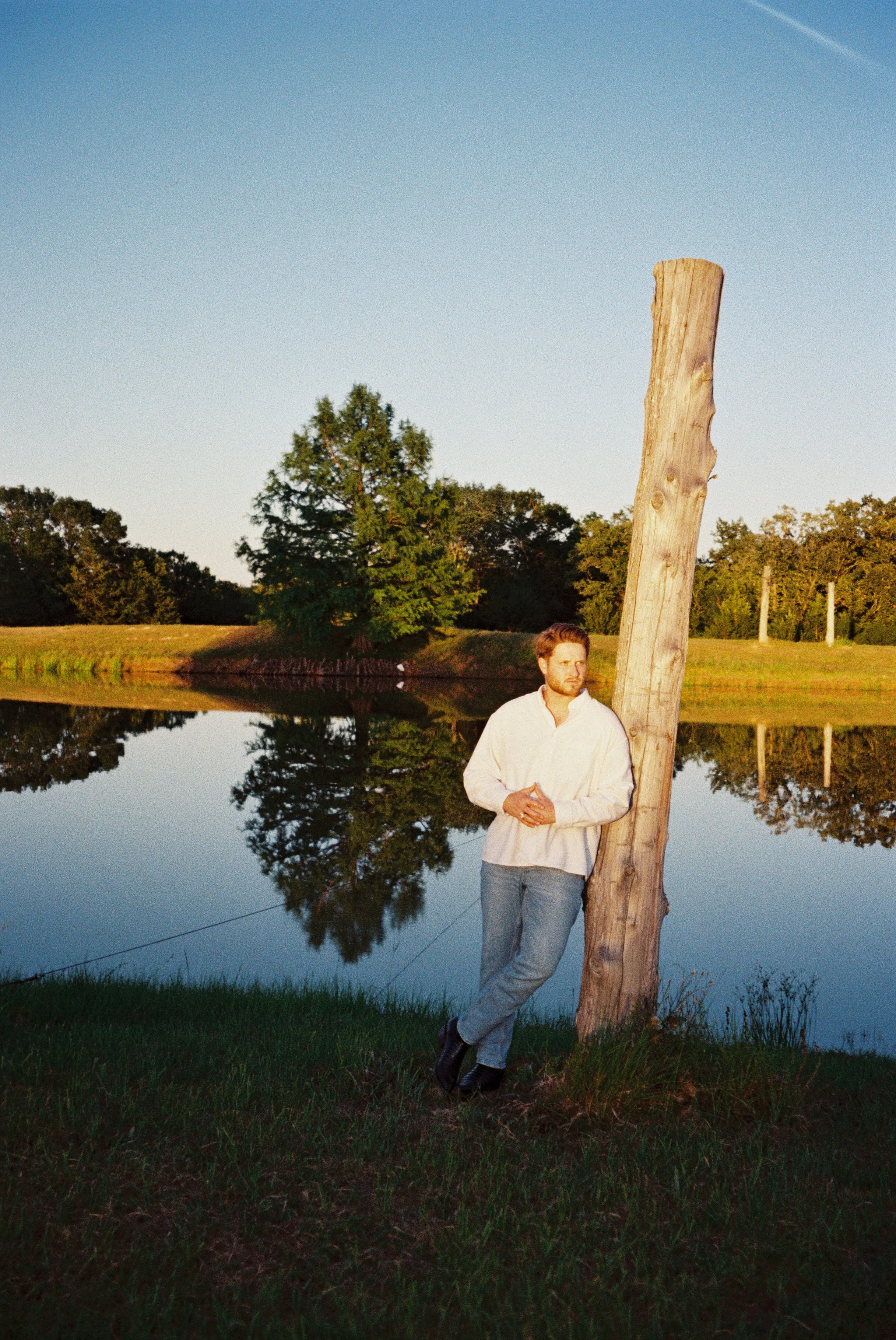 A man standing outdoors near a calm body of water with trees in the background, leaning on a tall, weathered wooden post, dressed in a light-colored shirt and jeans, during late evening or early morning.