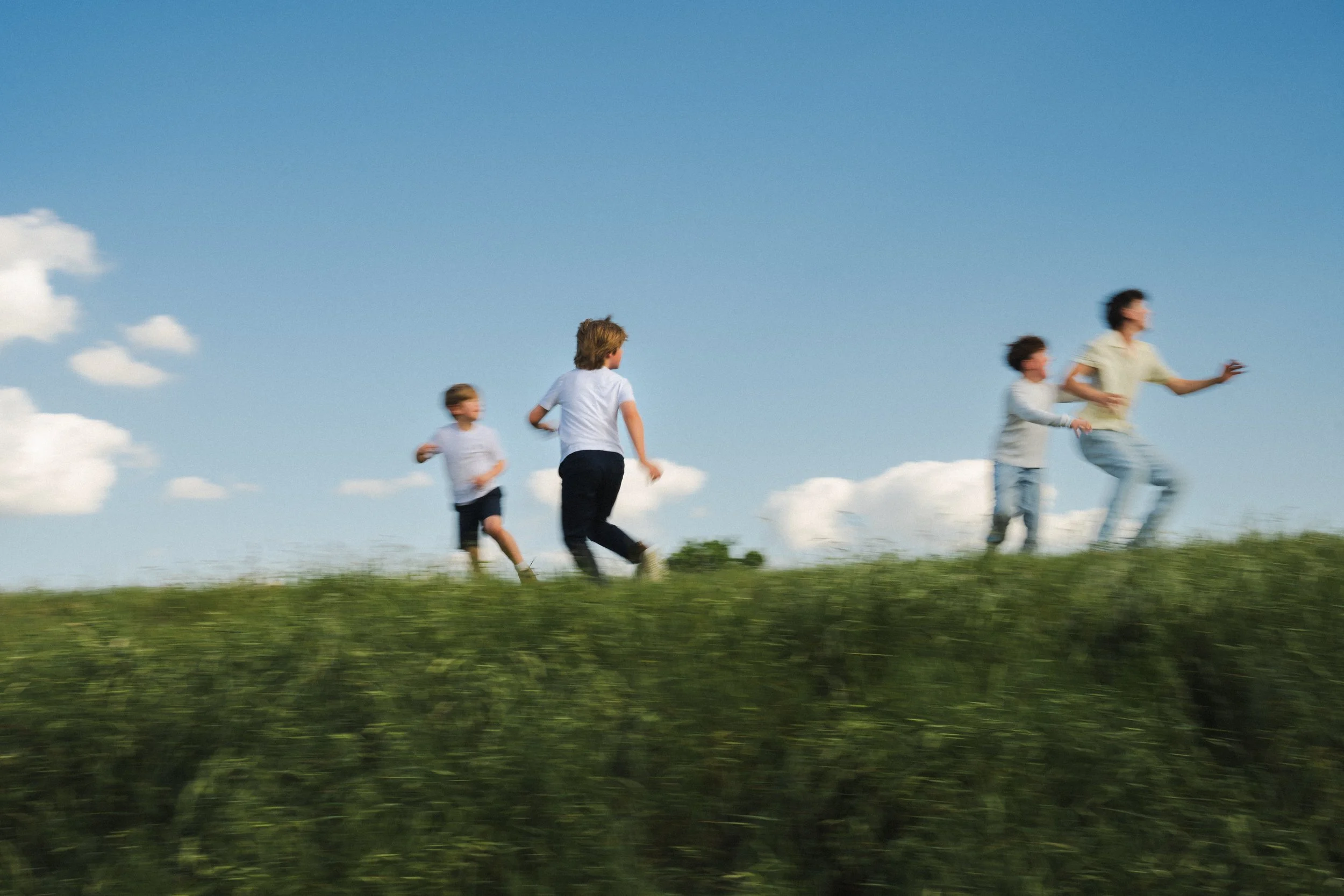 Children and an adult running on a grassy hill under a blue sky with white clouds.