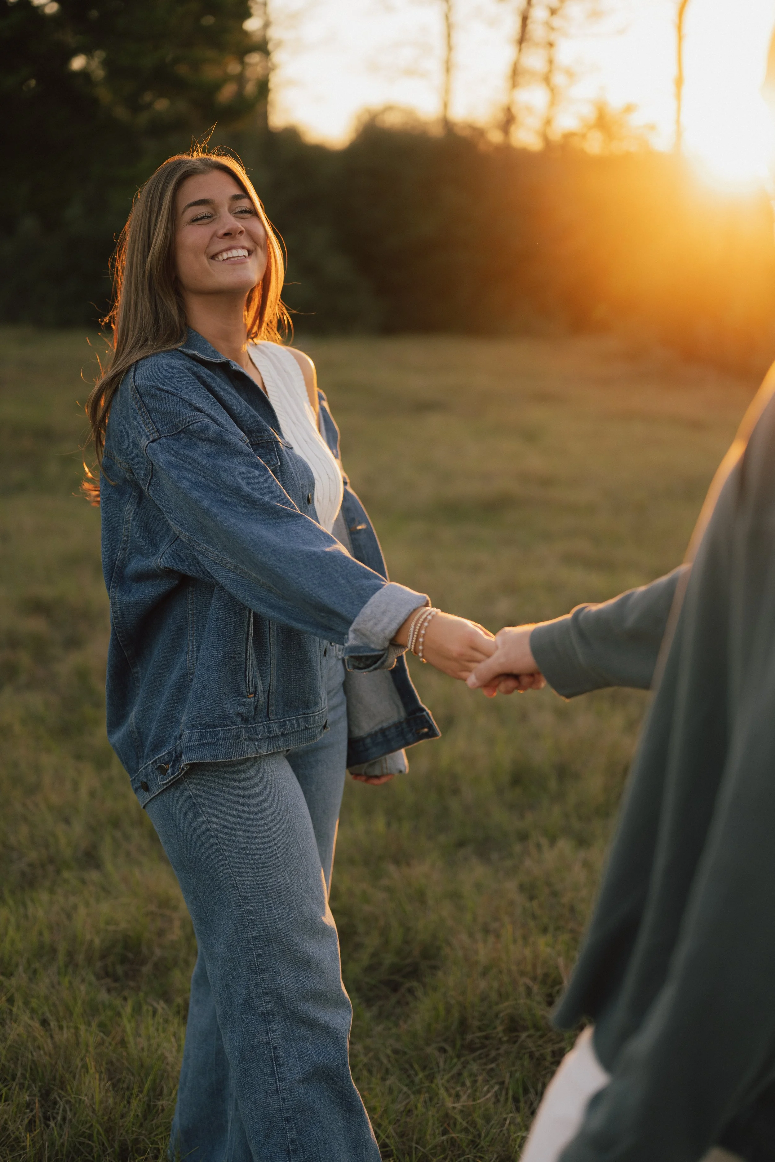 A smiling woman wearing a denim jacket shaking hands with someone outdoors at sunset.