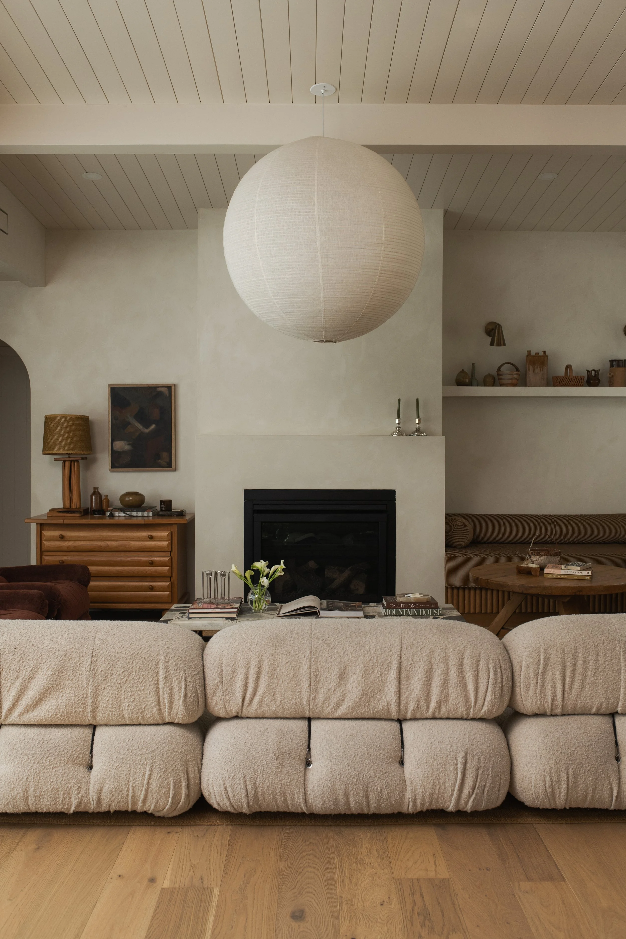A cozy living room with a beige sectional sofa, a wooden coffee table with books and a flower in a vase, a fireplace, and decorative items on shelves. A large paper lantern hangs from the ceiling, and there is a wooden dresser and wall art.