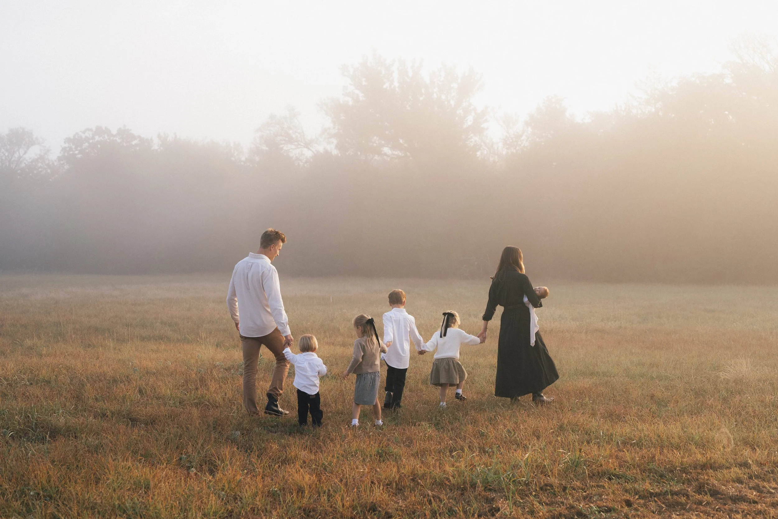 A family holding hands and walking through a field at sunset, with trees in the background.