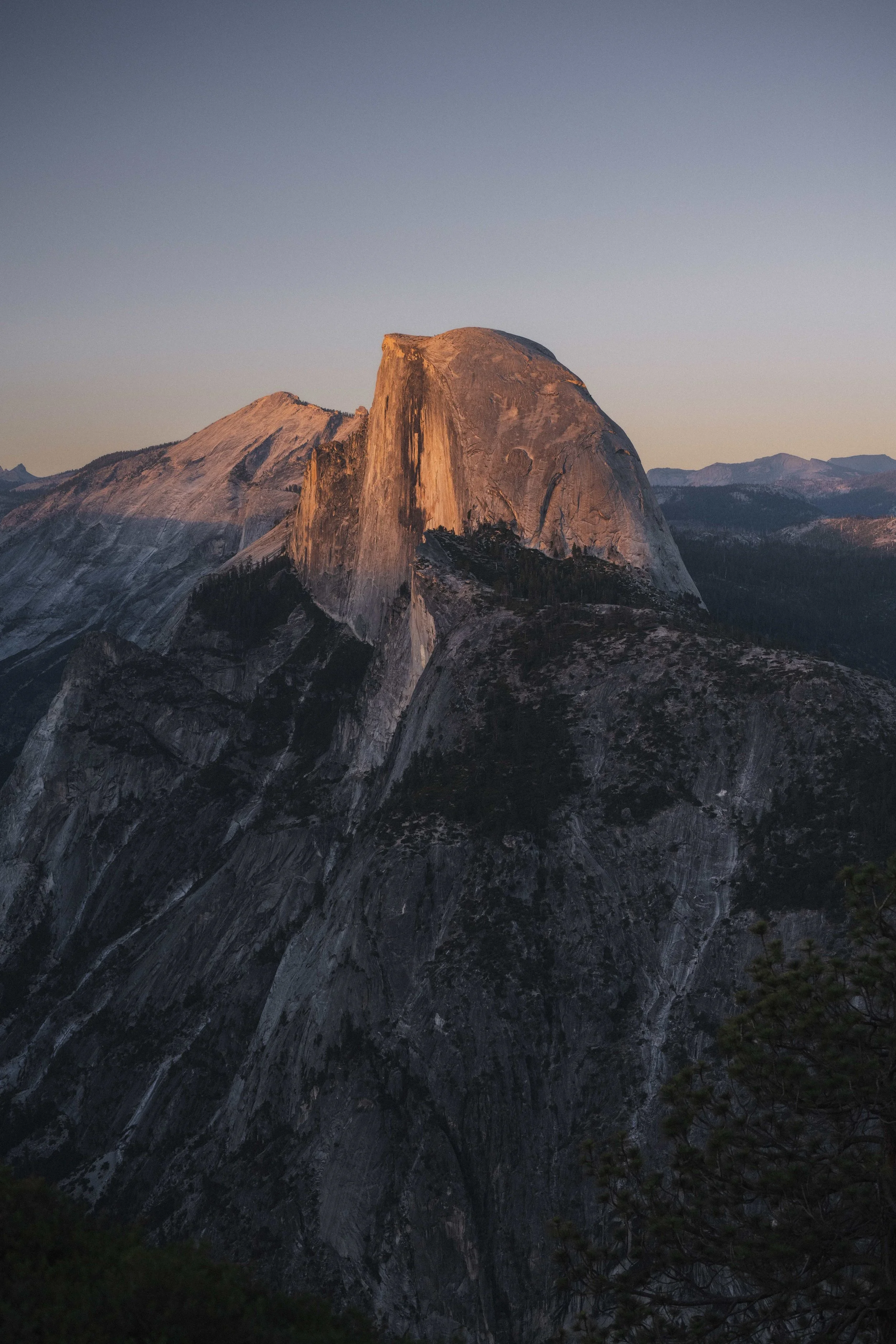 A landscape view of the Half Dome mountain in Yosemite National Park during sunset, with clear sky and shadows on the rocky surface.