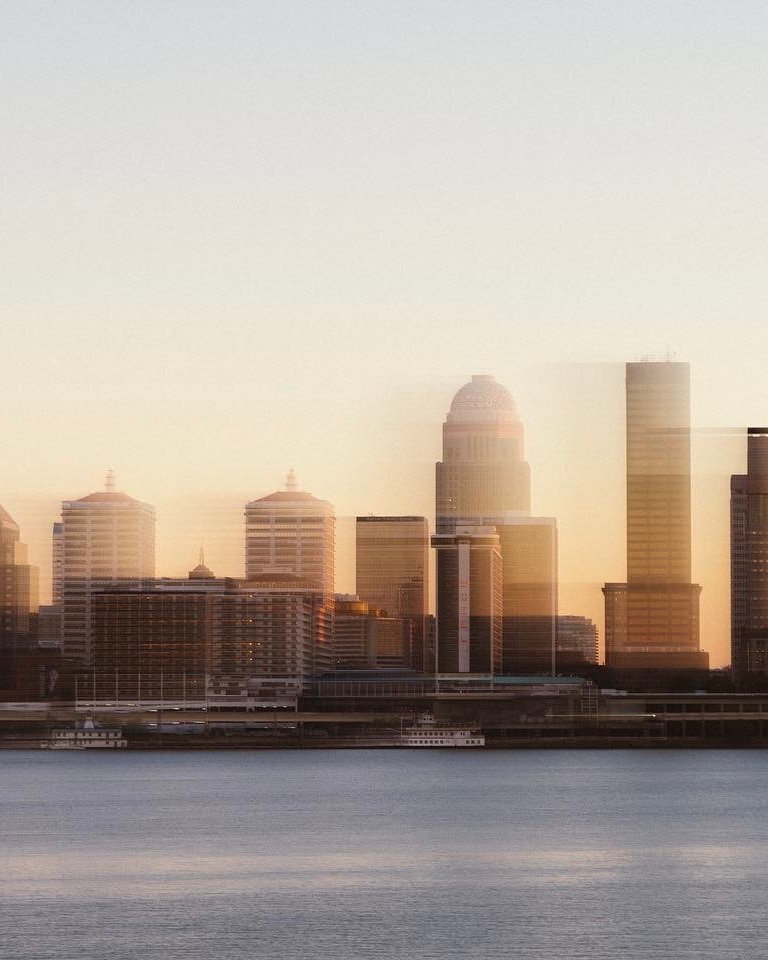 Faded city skyline with tall buildings and a body of water in the foreground, during sunset or sunrise.