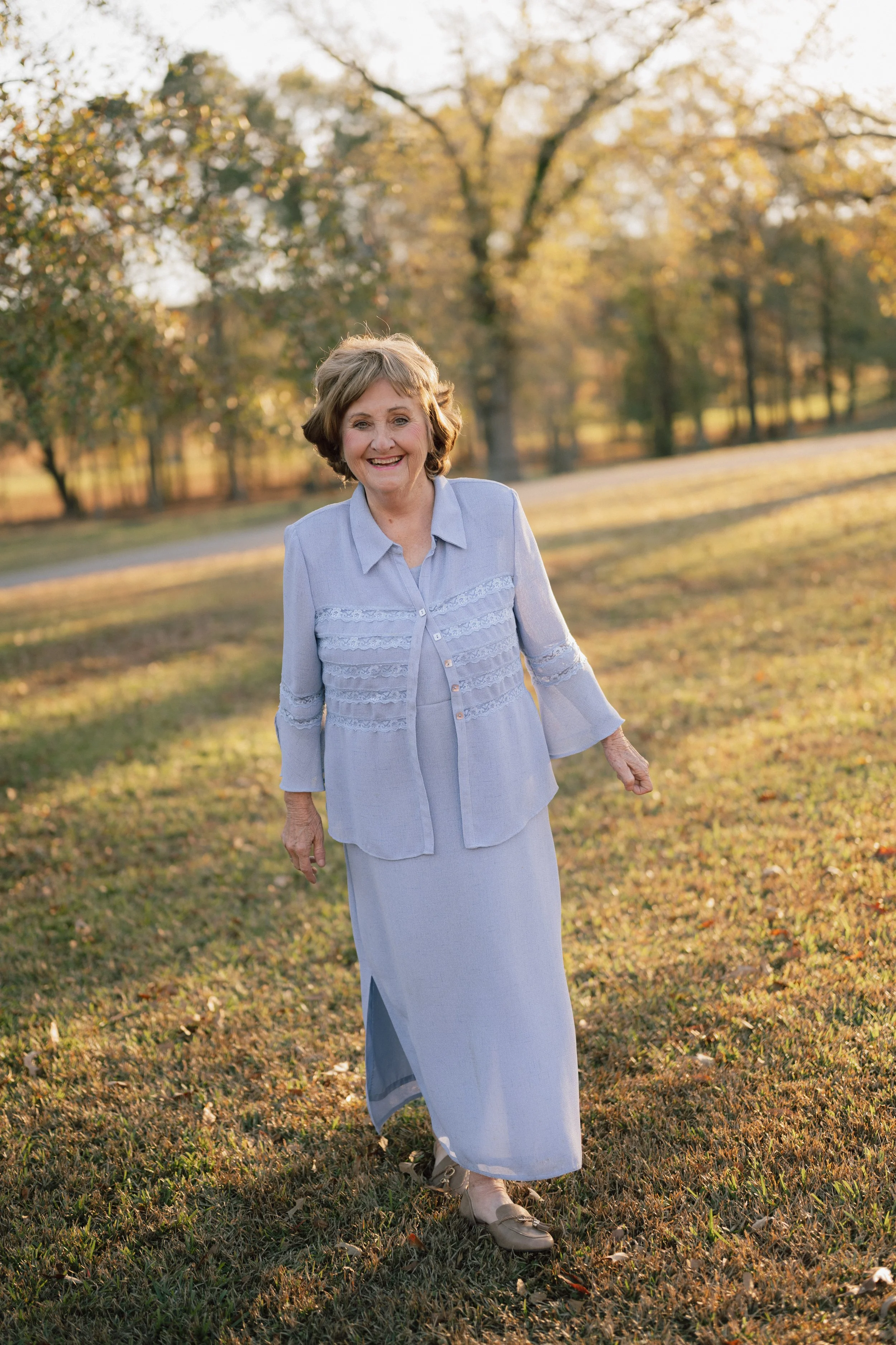 An elderly woman in a light blue dress and jacket walking on grass in a park during autumn, with trees in the background and sunlight filtering through the leaves.