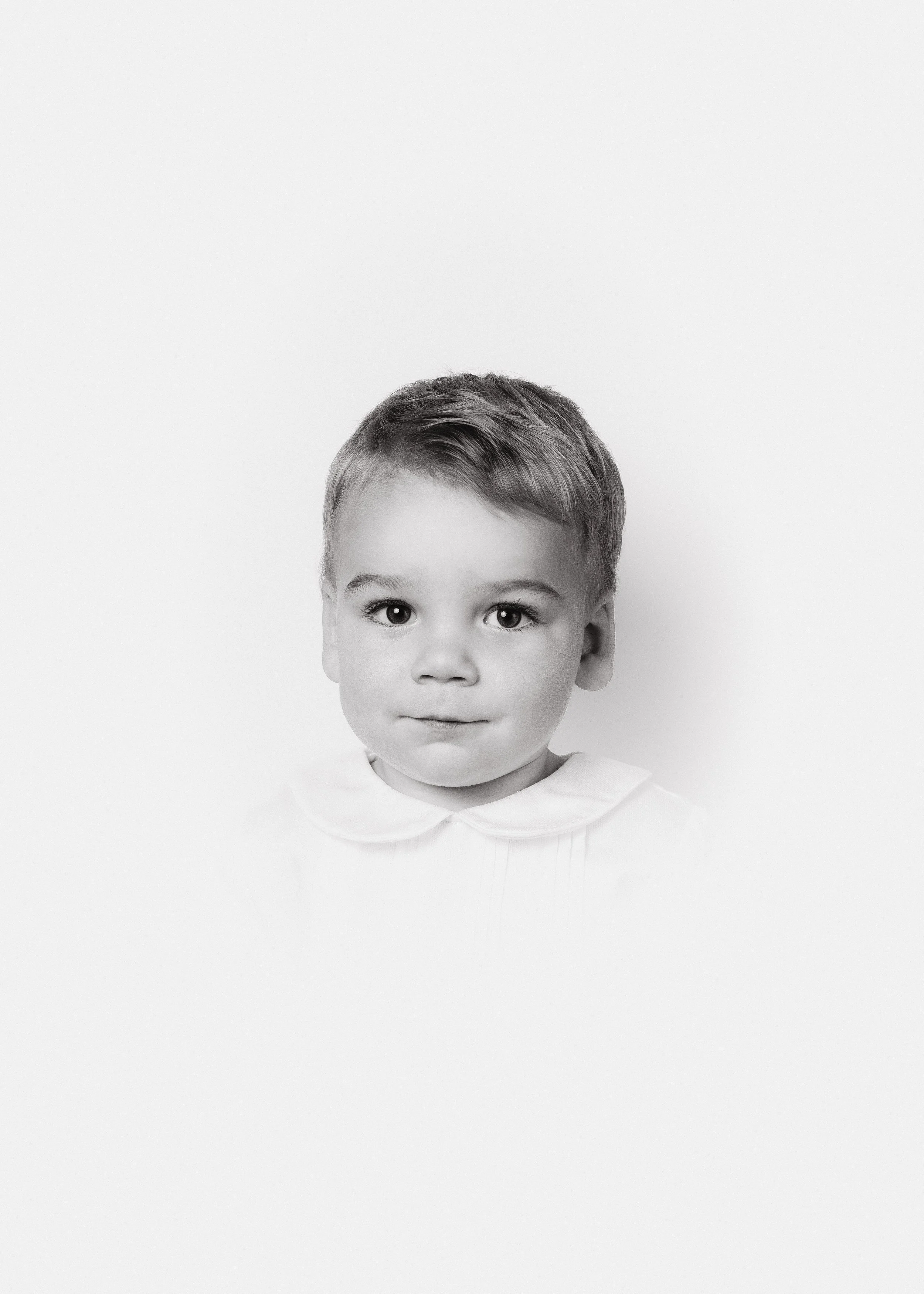 Black and white portrait of a young boy with short hair, wearing a collared shirt, looking directly at the camera with a neutral expression, against a plain white background.