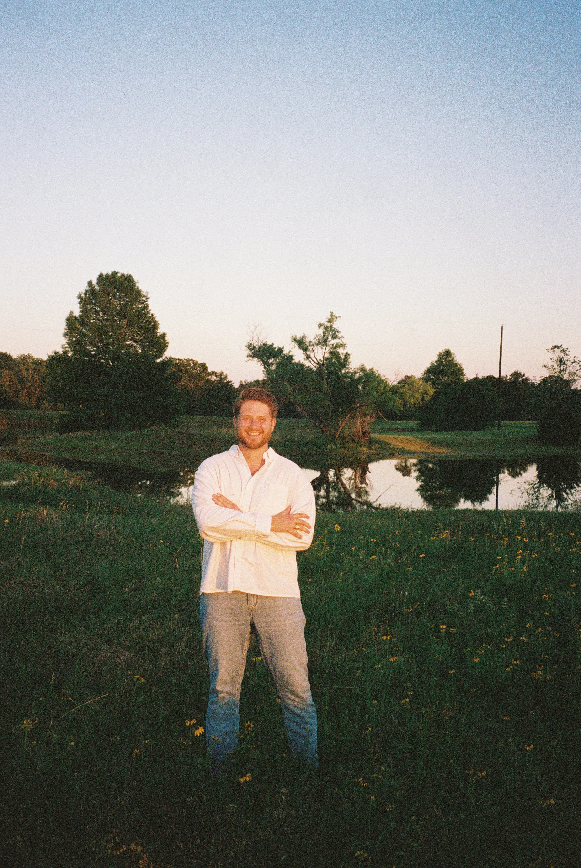A smiling man with a beard standing outdoors in front of a pond, trees, and a clear sky at sunset.