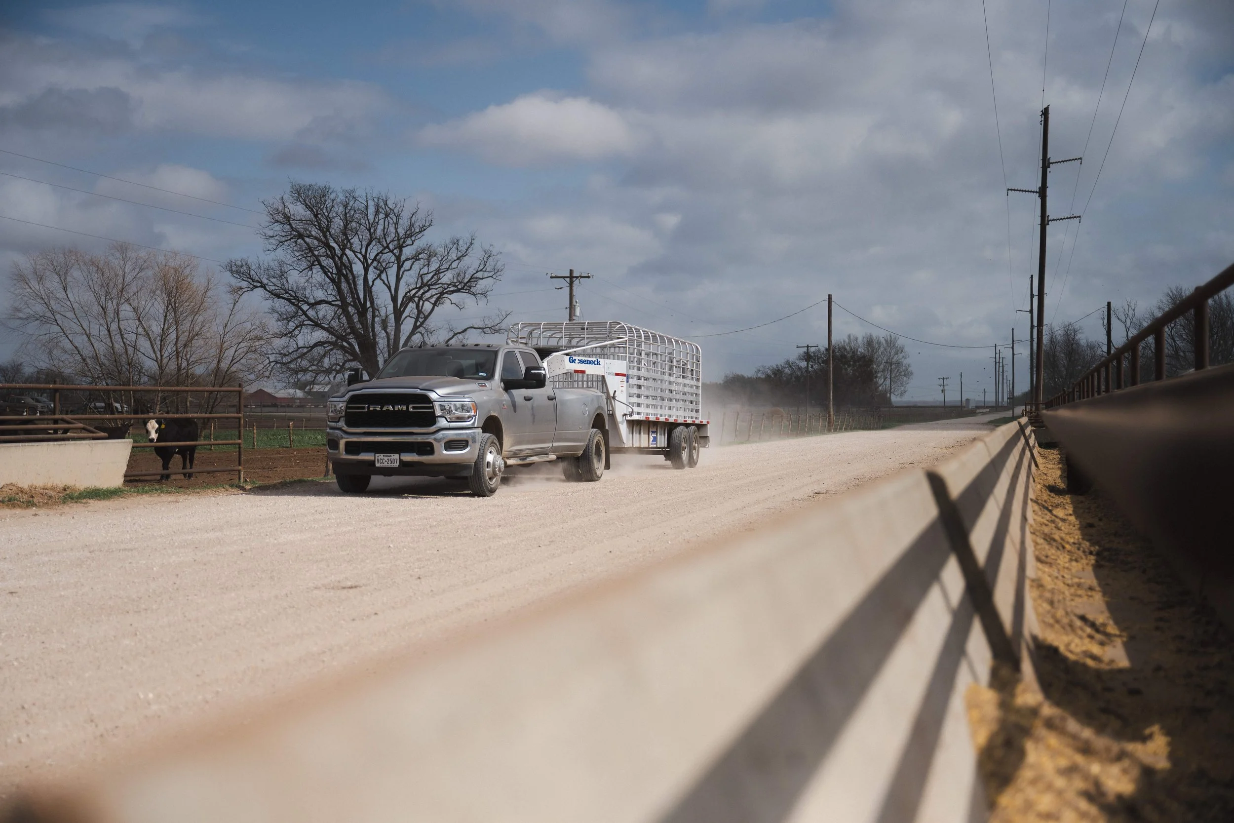 A gray pickup truck driving on a dirt road in a rural area with leafless trees, utility poles, and cloudy sky in the background.