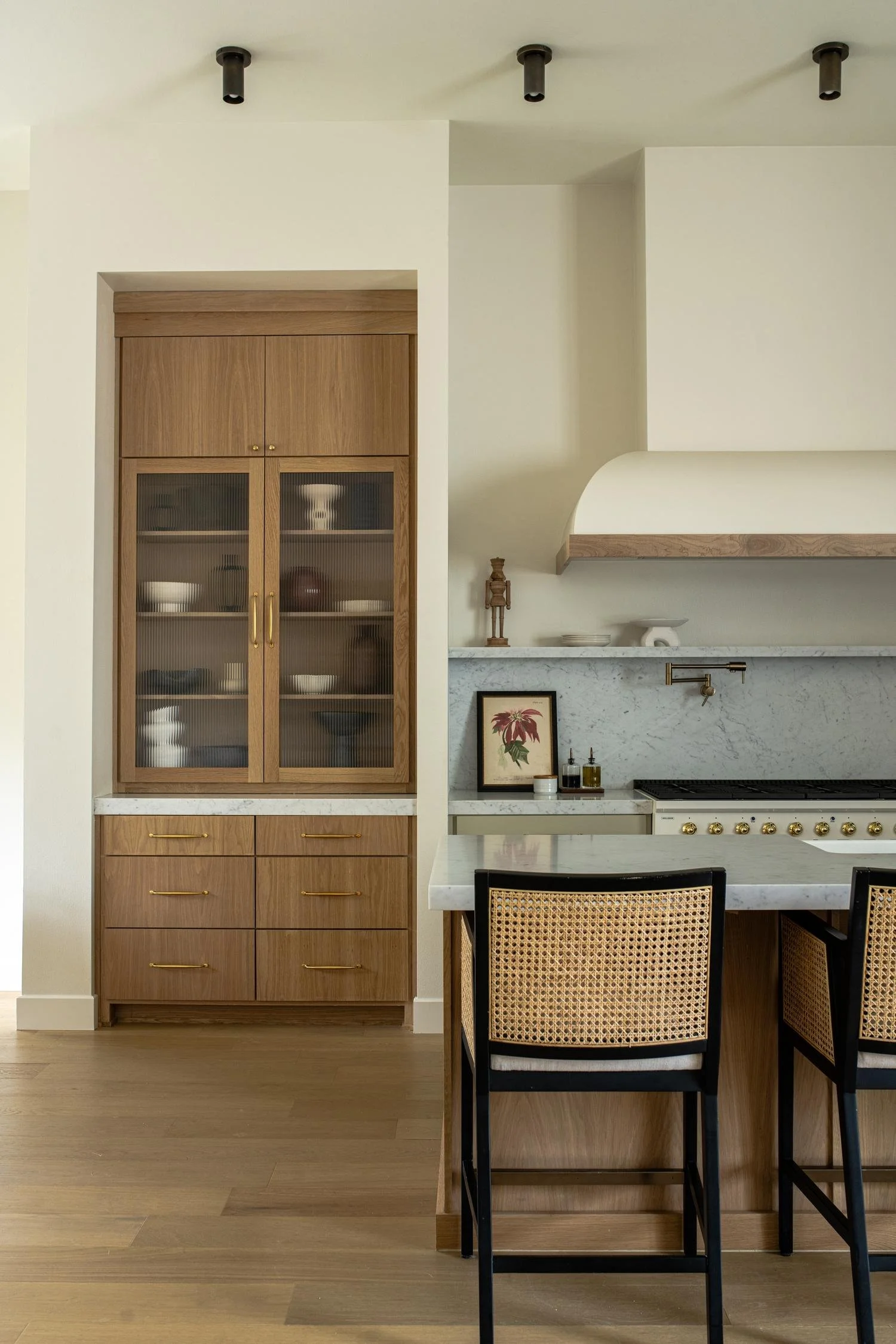 A kitchen with light walls and wooden flooring, featuring a tall wooden cabinet with glass doors displaying dishes, and a kitchen island with black chairs that have woven cane backrests, a white marble countertop, and a framed botanical print on the backsplash.