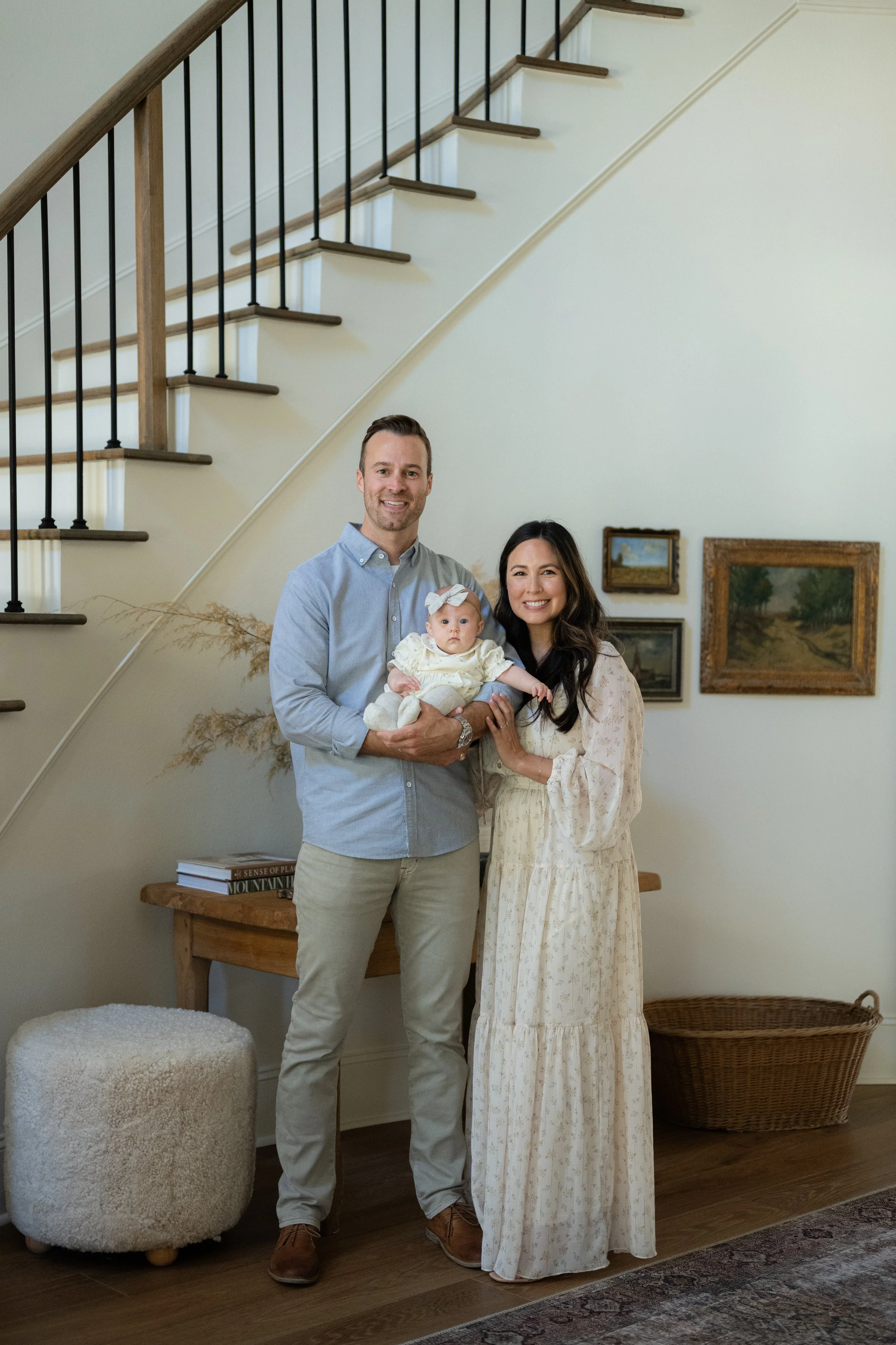 A family of three posing indoors, standing in front of a staircase. The man on the left holds a baby girl, and a woman stands beside them. The setting includes wall-mounted paintings, a side table, and a woven basket in the background.