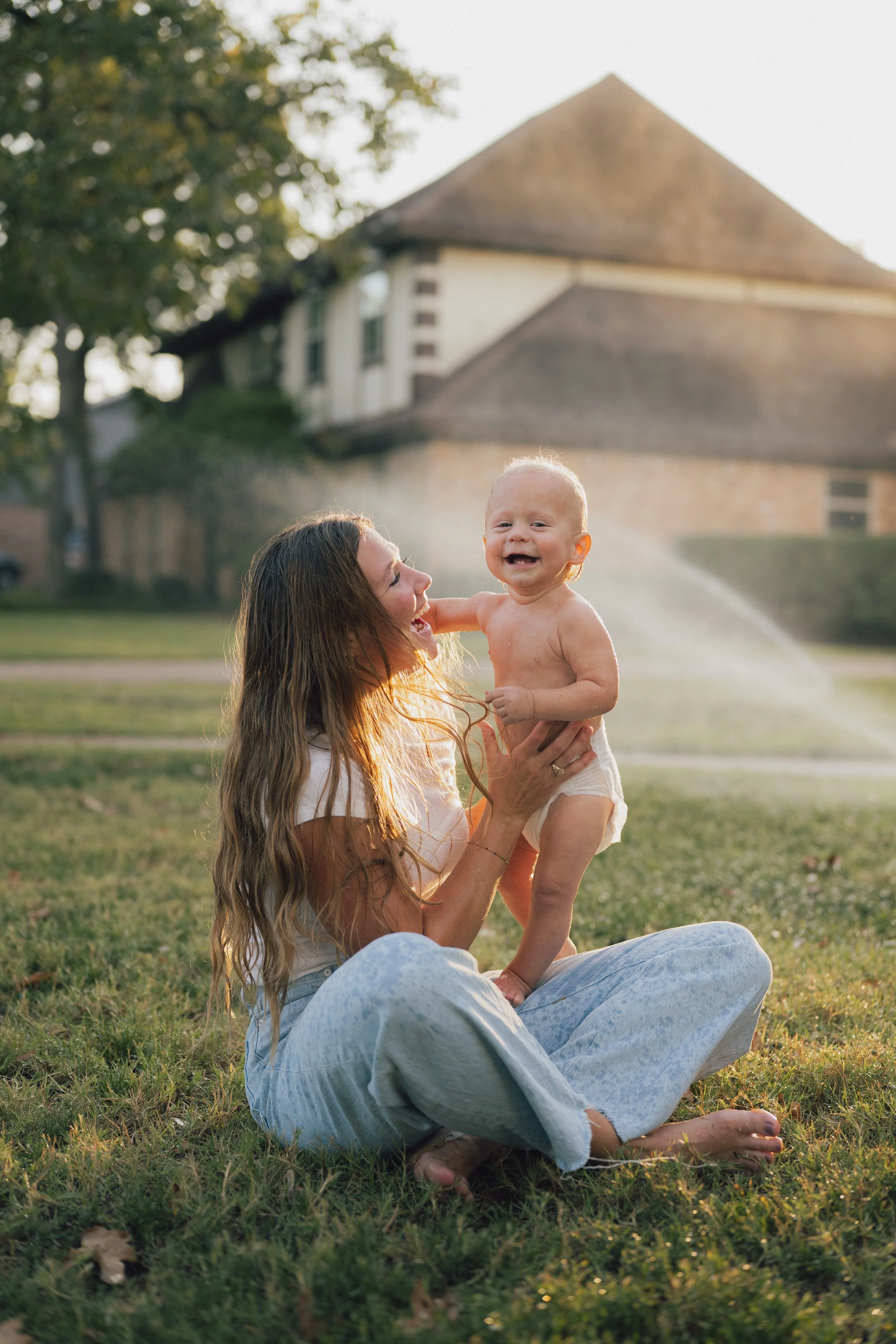 A woman with long hair sitting on grass, holding a smiling, shirtless toddler with curly hair against a backdrop of trees and a house with a sloped roof, during sunset.