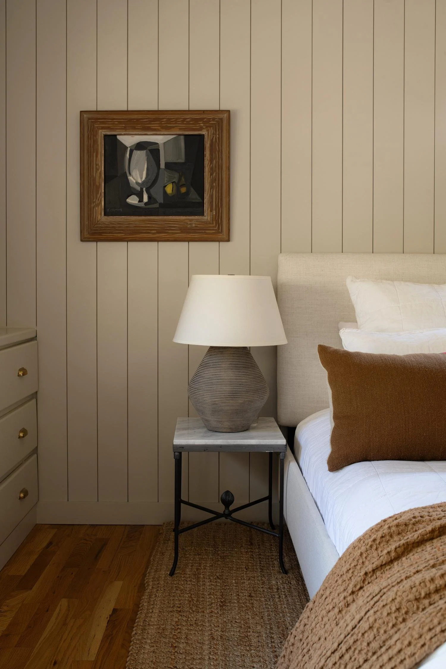 A bedroom corner with a white bed and brown pillow, wooden floor, a beige nightstand with a ceramic lamp, a framed abstract painting on a beige paneled wall, and a woven rug.