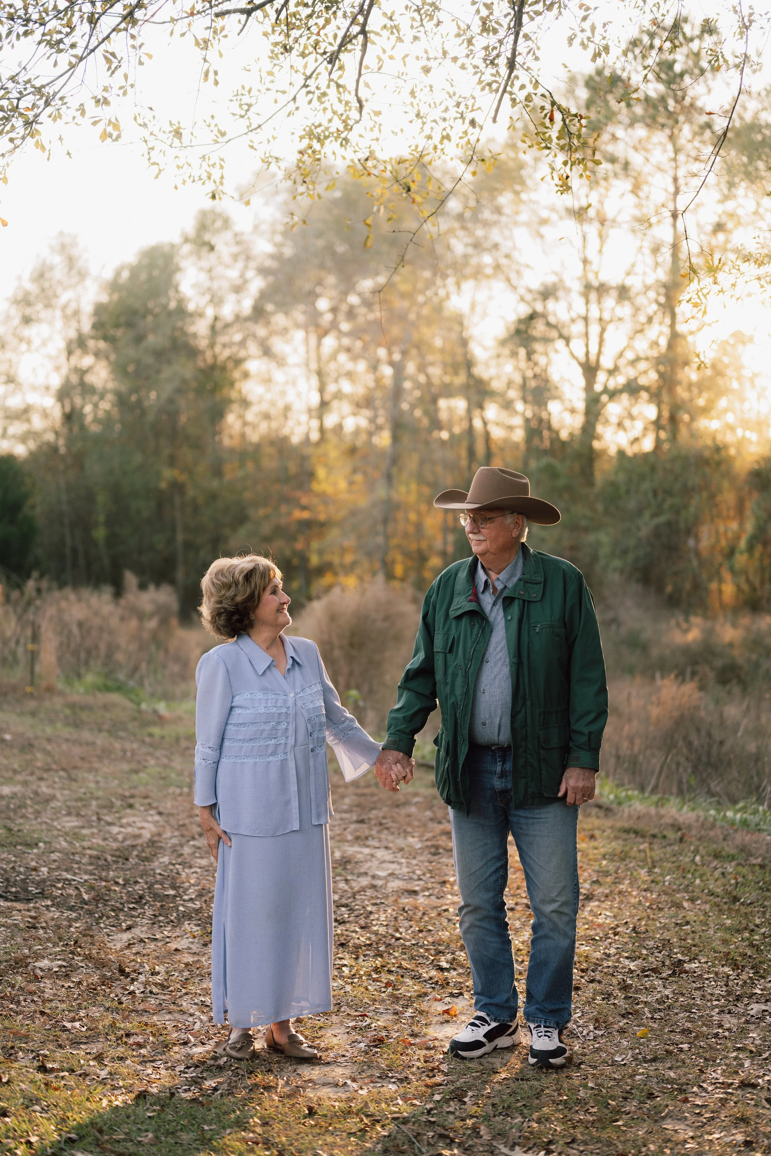 An elderly couple holding hands and walking outdoors on a fall day, with the woman looking up at the man who is wearing a wide-brimmed hat and glasses, in a natural setting with trees and sunlight.