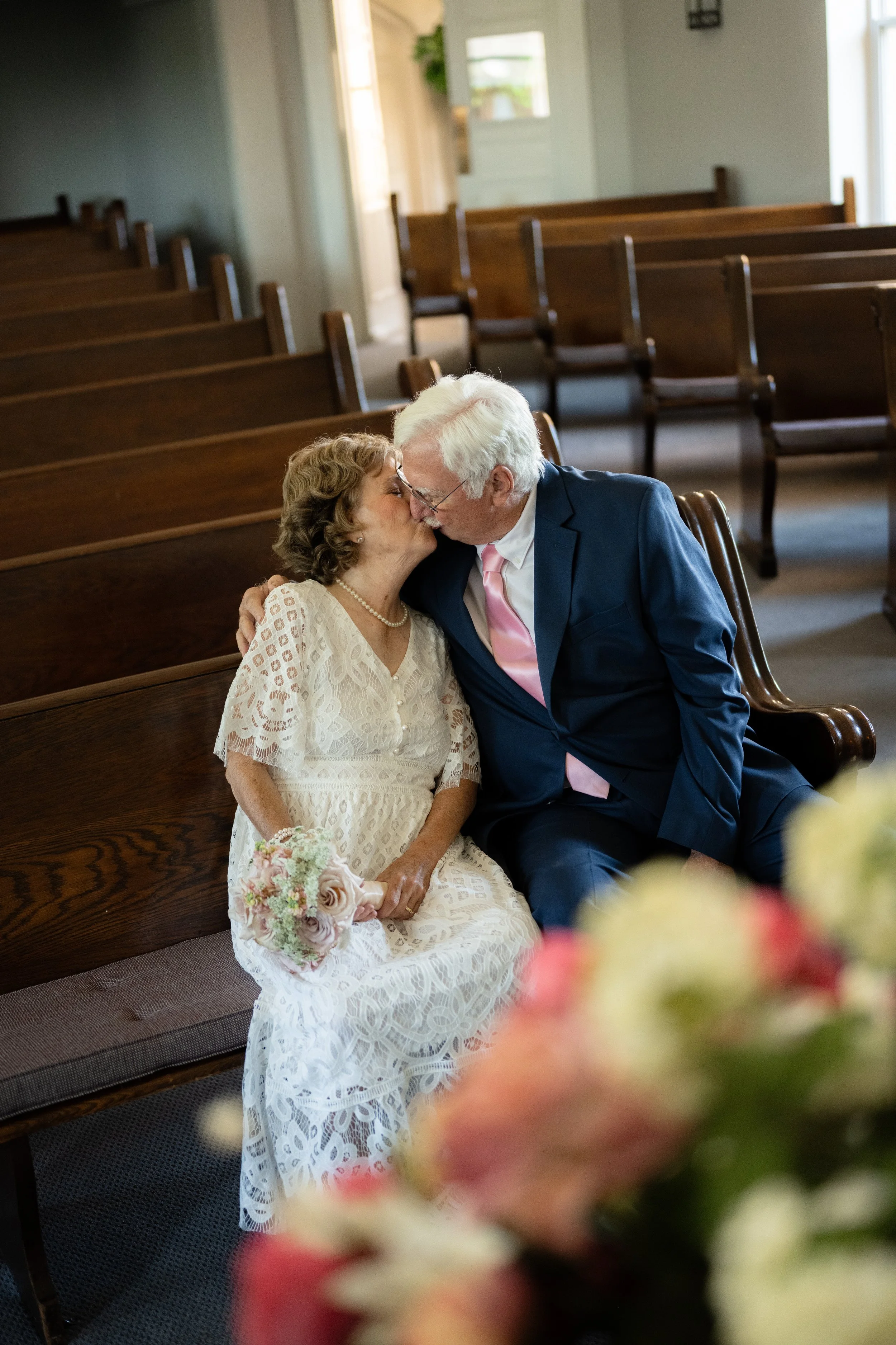 An elderly couple in wedding attire sharing a kiss and holding a bouquet of flowers inside a church with empty wooden pews.
