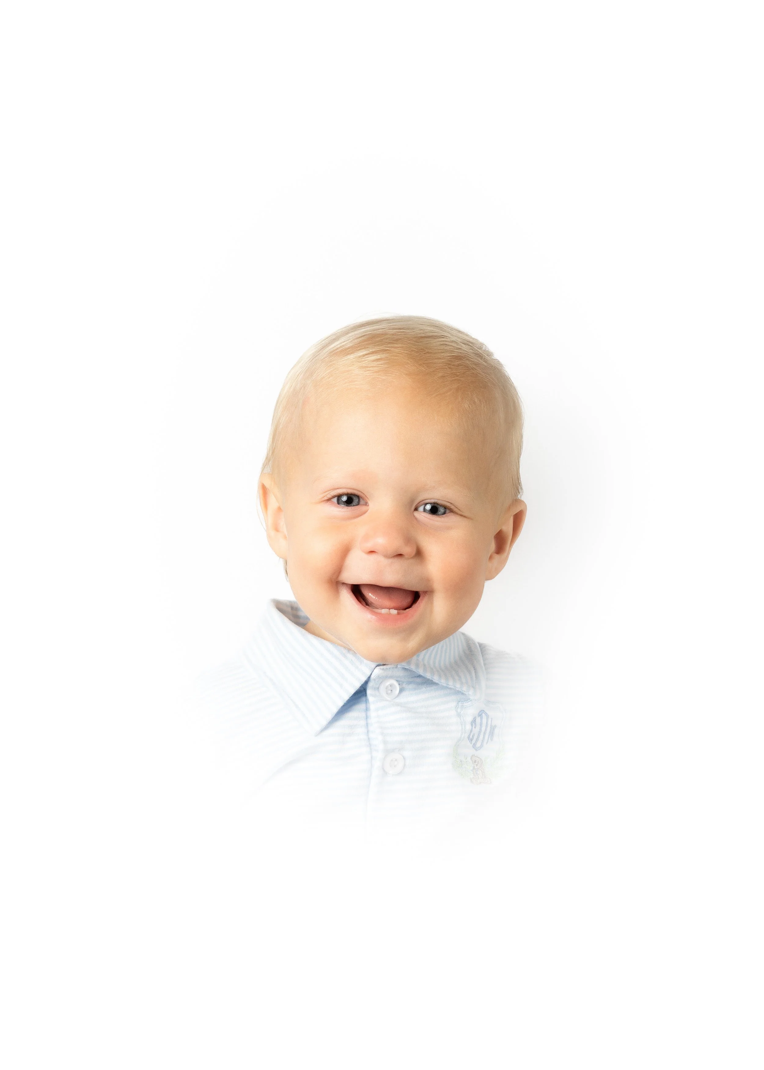A smiling young boy with blonde hair and blue eyes, wearing a light blue collared shirt, against a plain white background.