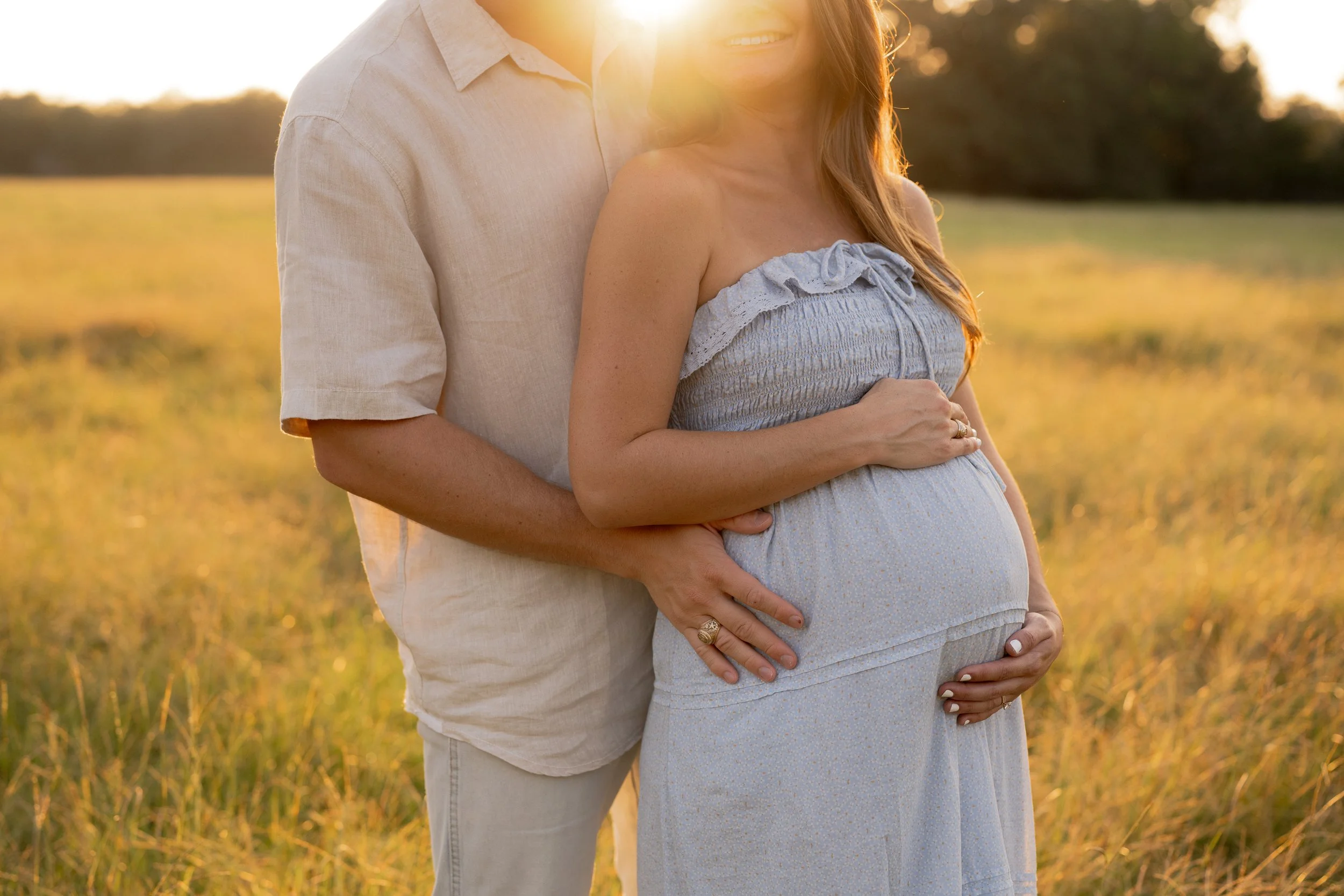 A pregnant woman in a light blue dress stands outdoors with a man in a light-colored shirt, both embracing during sunset in a grassy field.