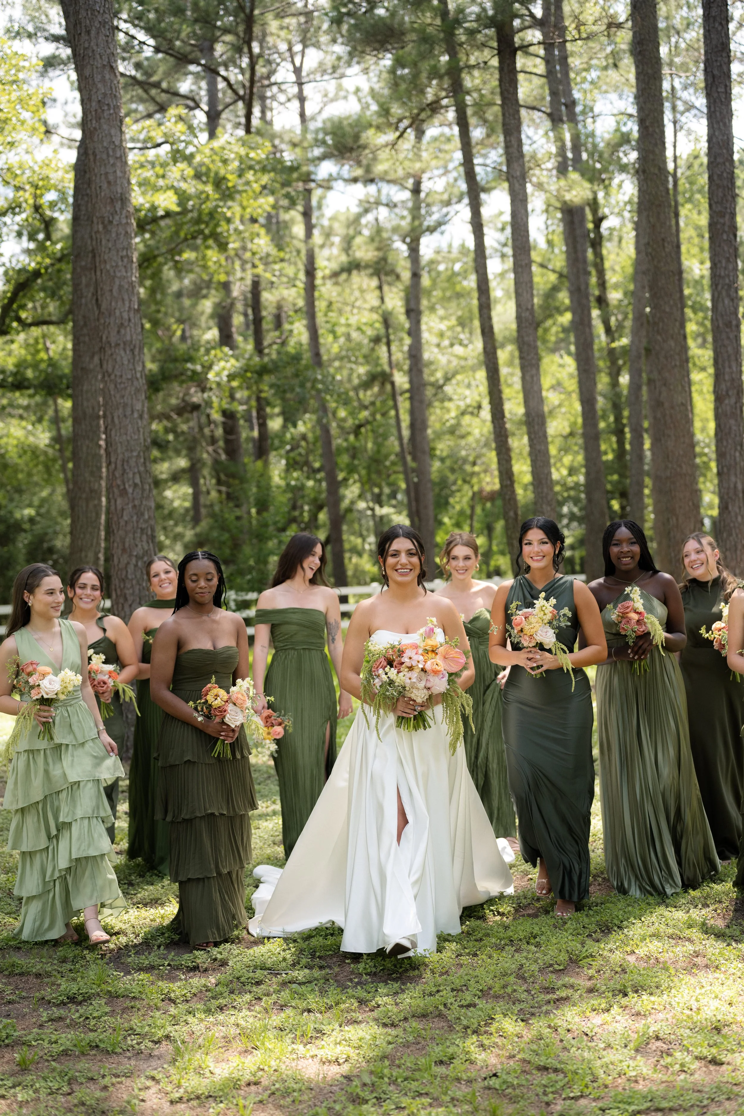 A group of women in green dresses walking outdoors in a forest setting, with the bride in a white gown leading the way, smiling and holding a bouquet of flowers.