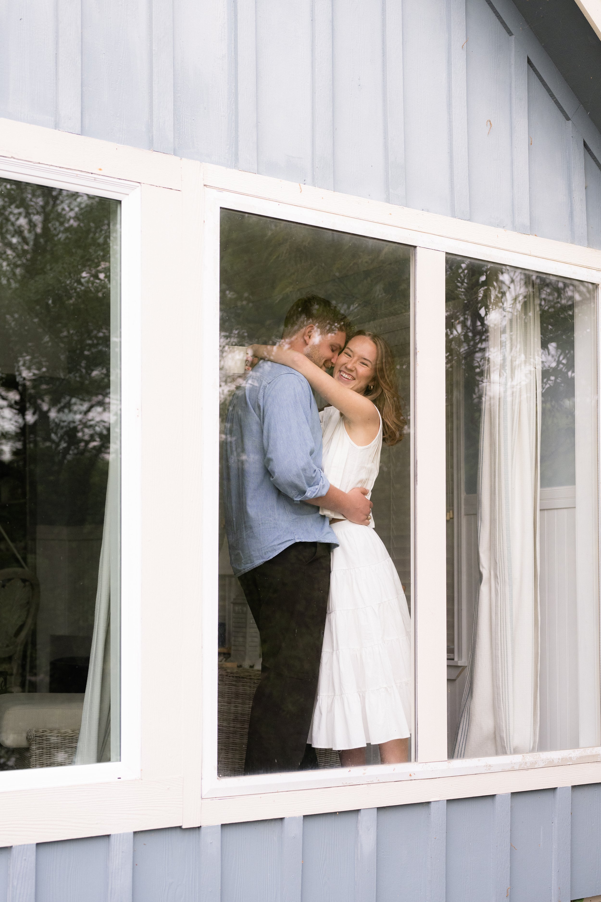 A couple hugging inside their home, viewed through a large window from the outside. The woman is smiling, and the man appears to be whispering to her.