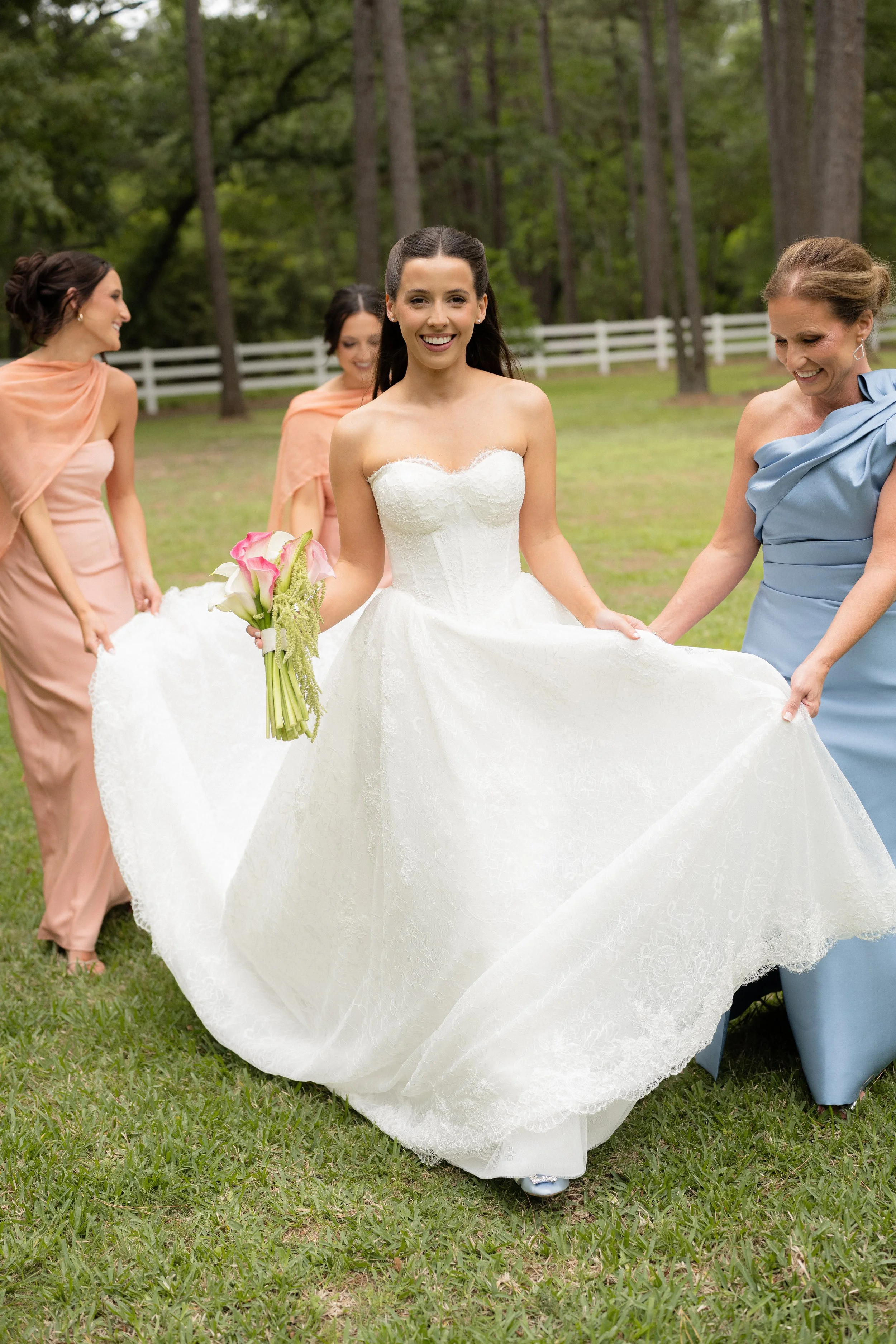 A bride in a strapless white wedding dress holding a bouquet of pink and white calla lilies, smiling, surrounded by bridesmaids in peach and blue dresses, outdoors with tall trees and a white fence in the background.