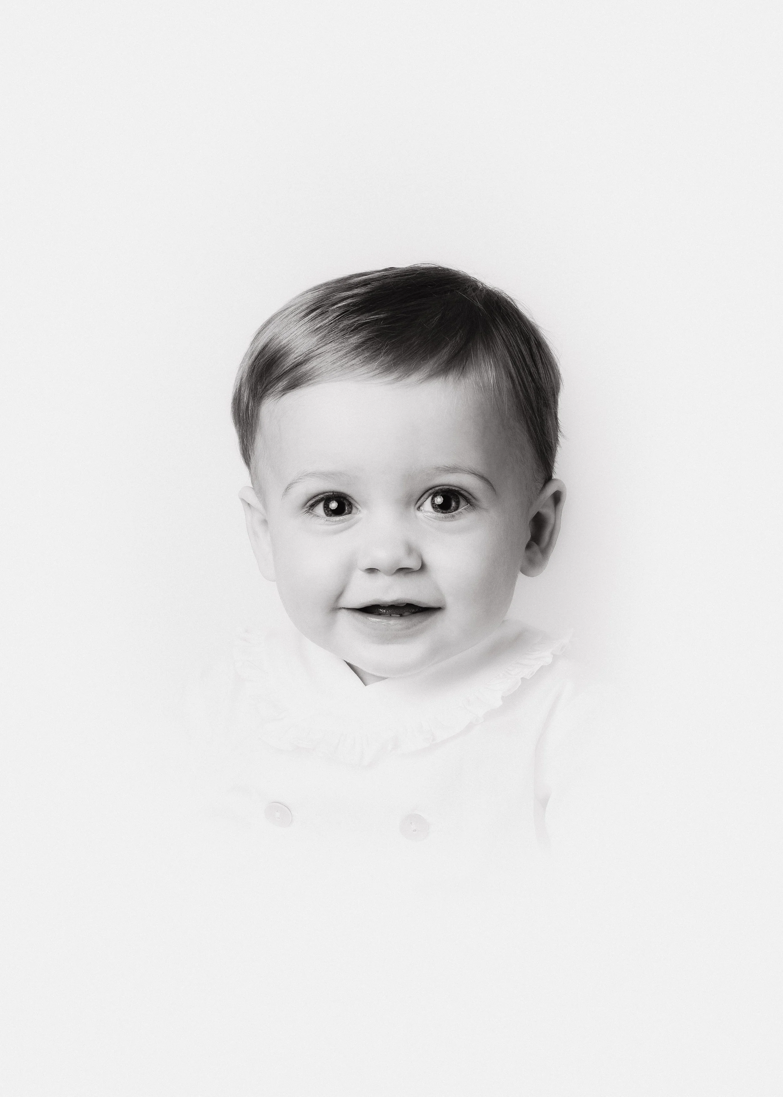 Black and white portrait of a young child with short hair, smiling, wearing a light-colored top with a ruffled collar.