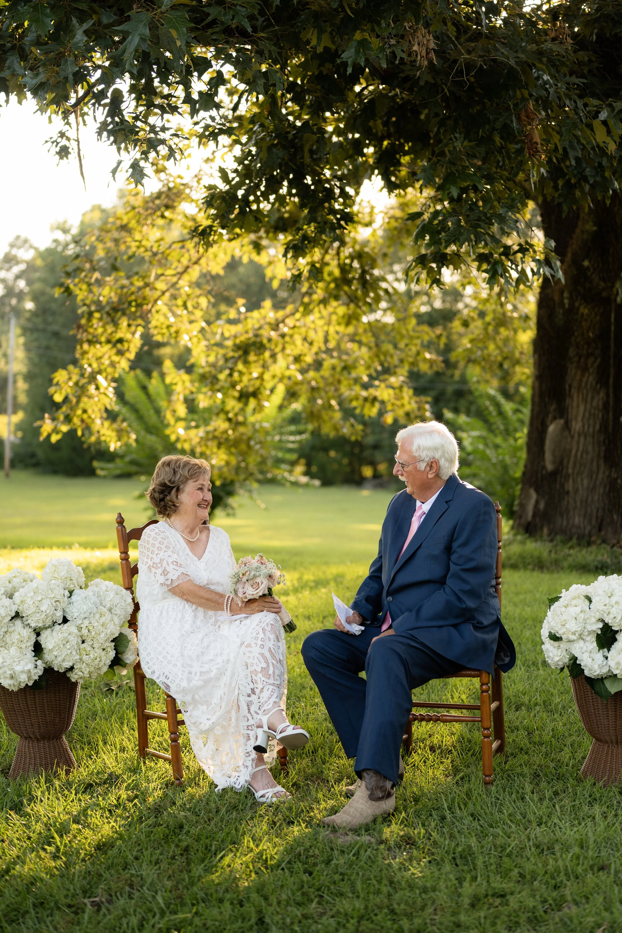 An elderly woman and man are sitting outdoors on wooden chairs during a wedding ceremony. The woman holds a bouquet of flowers and wears a white dress, while the man wears a navy suit and pink tie. They are smiling at each other as they participate in the event, surrounded by white floral arrangements on wicker baskets, with lush green grass and large trees in the background.