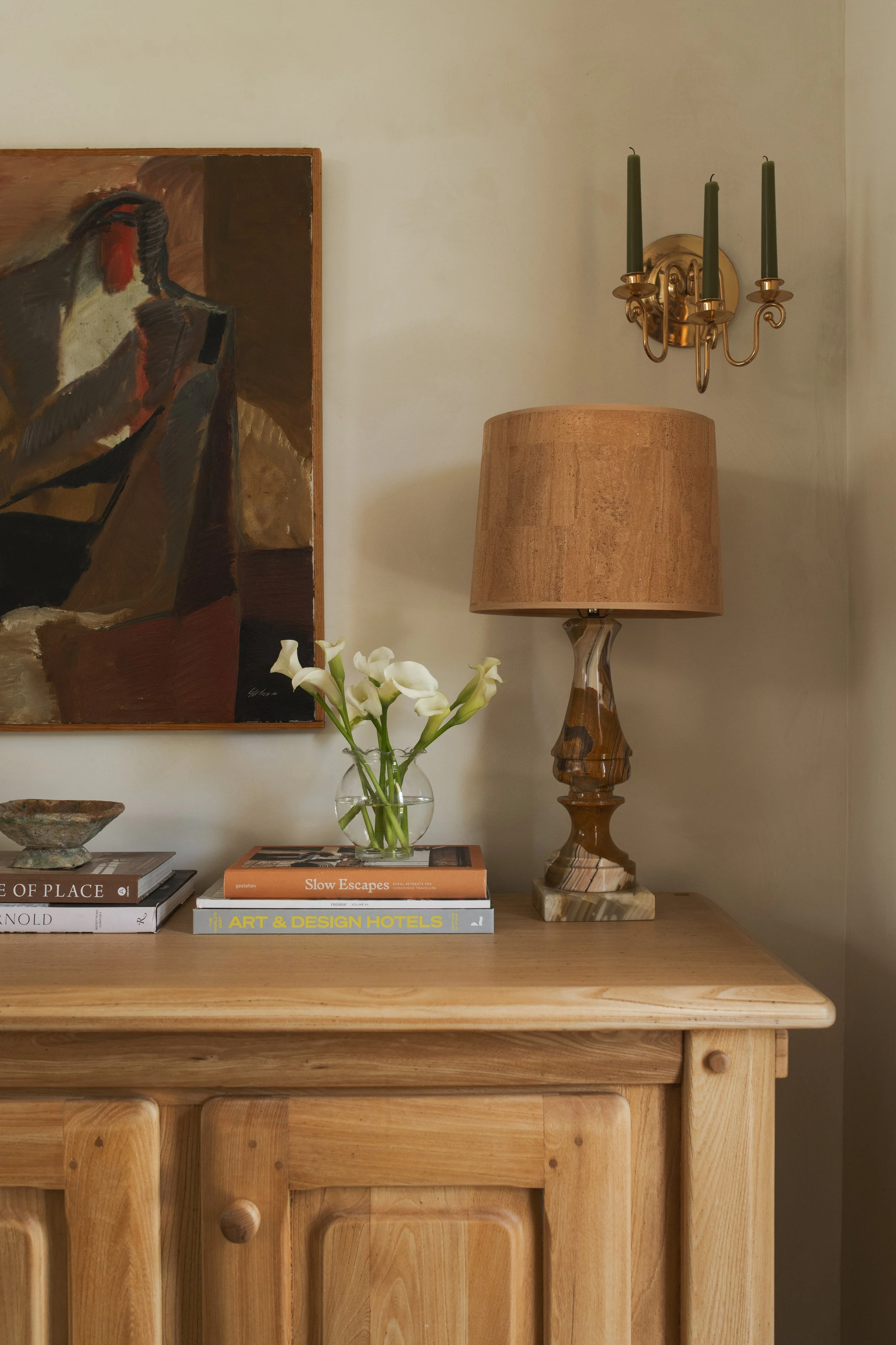 A wooden sideboard with a stack of three books, a clear glass vase with white calla lilies, a lampshade, a painting of a bird, and a brass wall-mounted candle sconce with three green candles.