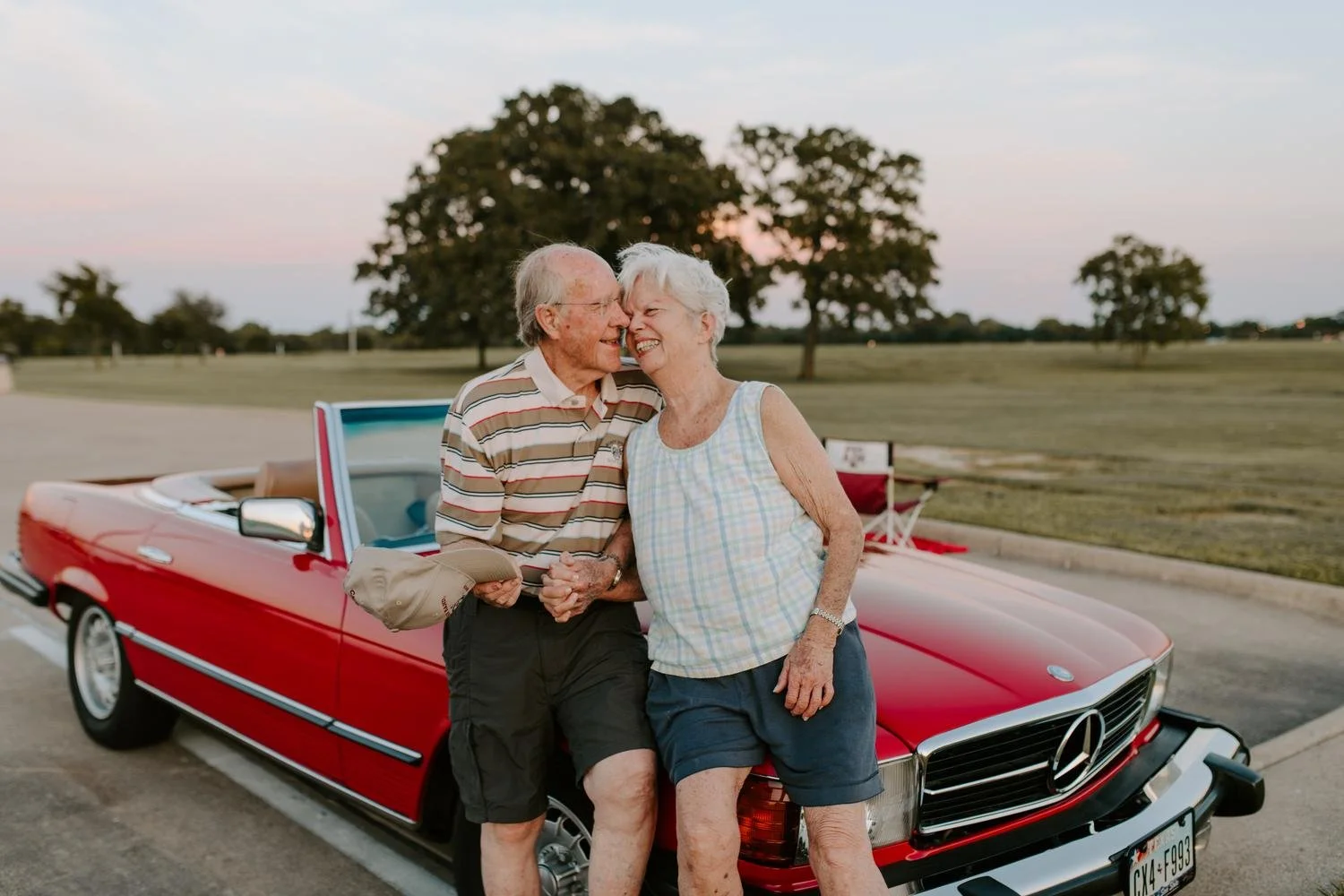 An elderly couple sharing a joyful moment, standing next to a red vintage convertible car in an open park at sunset.