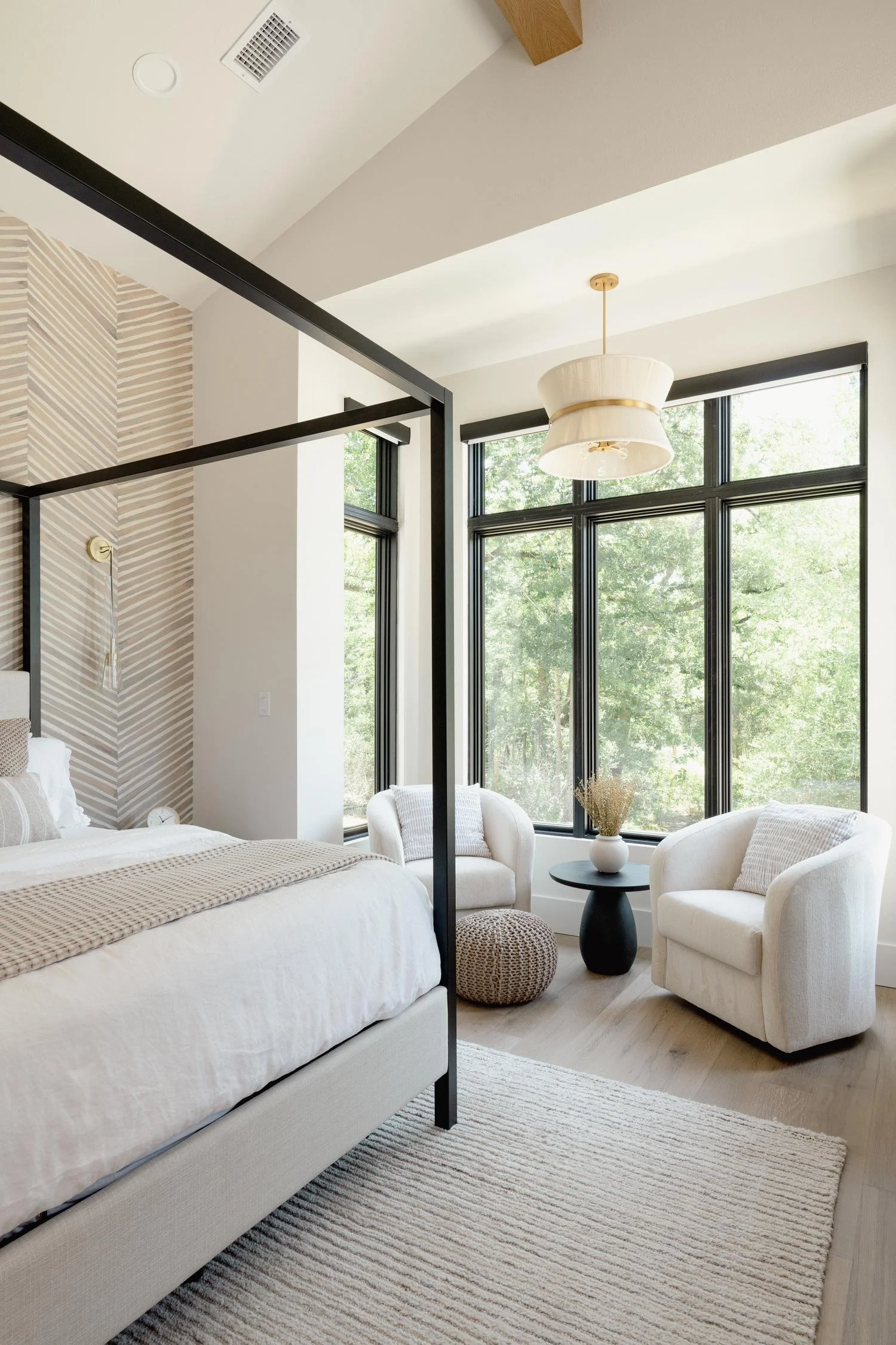 A bright bedroom with large windows, white and beige furniture, a beige woven rug, and a pendant light fixture. The bed has white bedding with striped pillows, and there are two white armchairs with a black side table and a decorative vase with dried