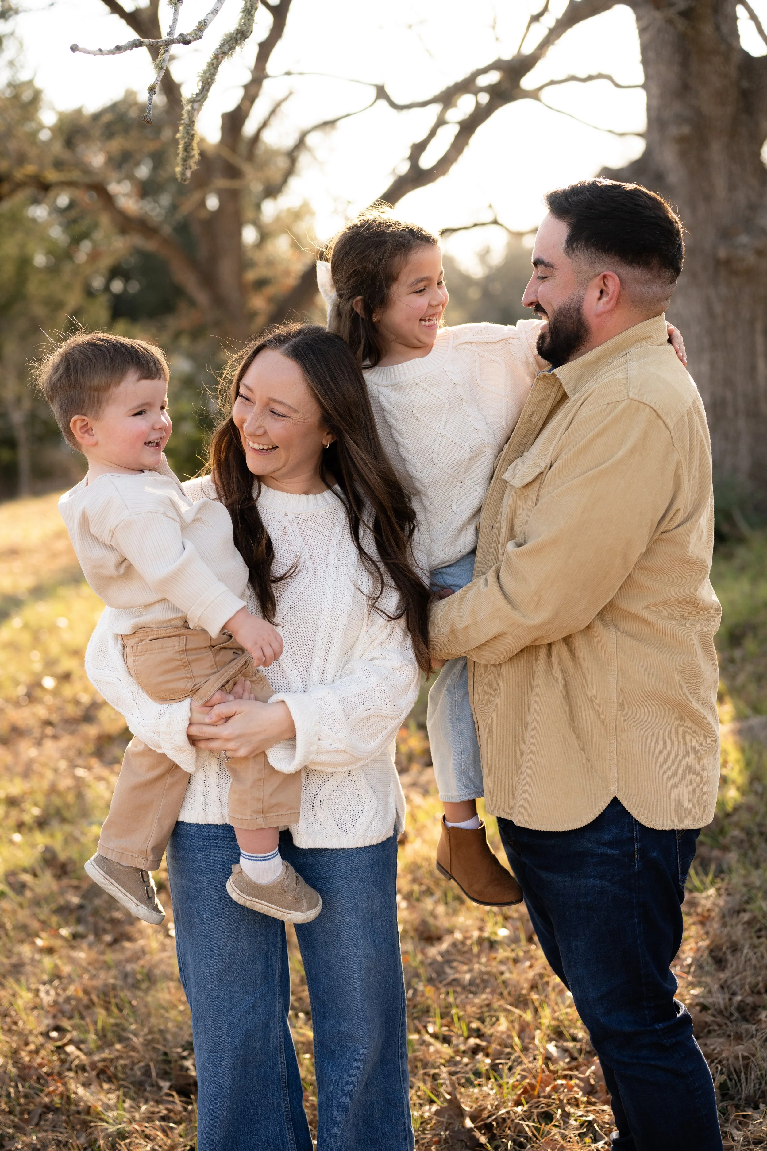 Family of four outdoors, smiling and laughing, during sunset, with trees and fallen leaves in the background.