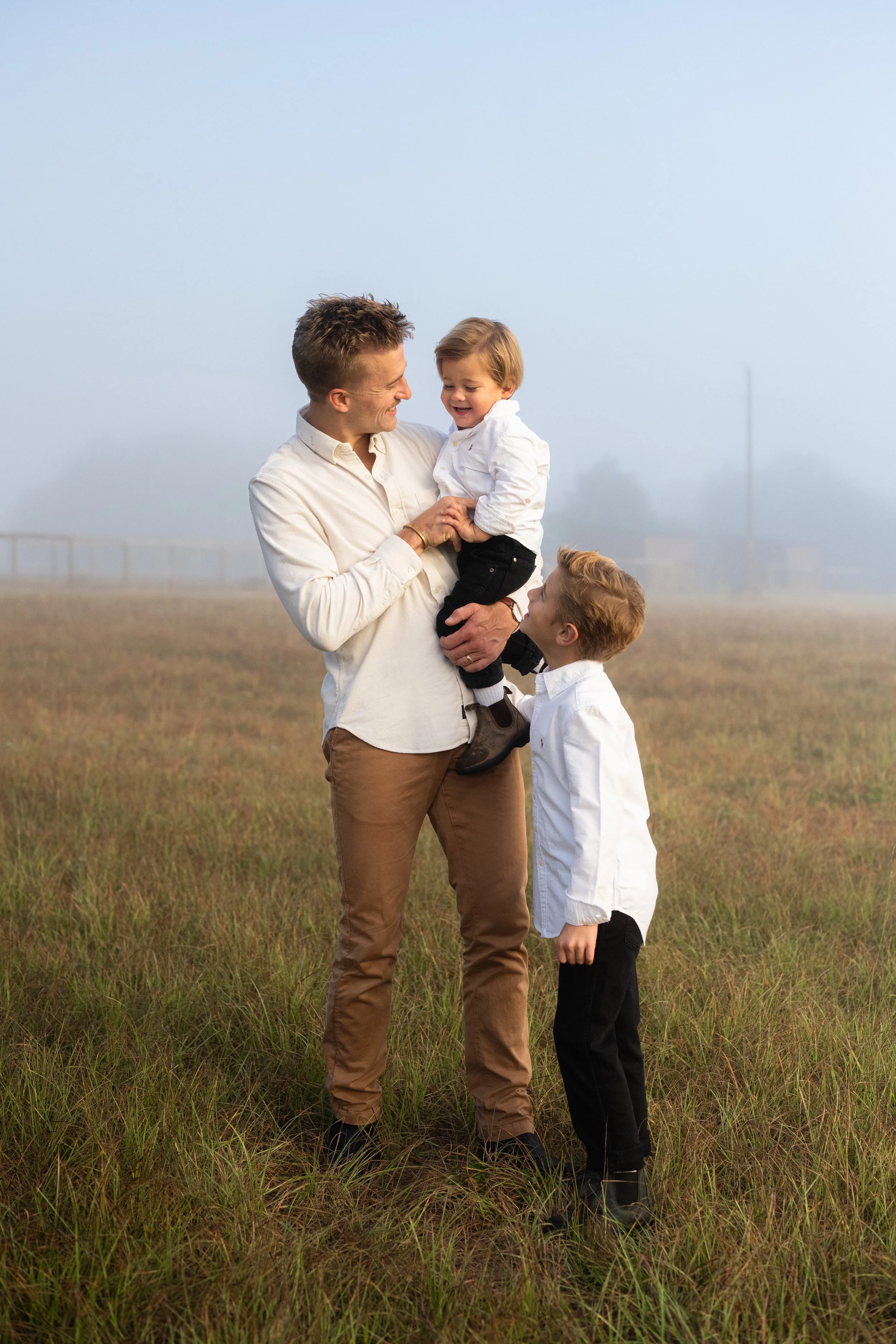 A man holding a young child in his arms while standing in a grassy field, smiling at each other. Another child stands nearby, looking up at the man. The background features a foggy landscape with a distant bridge.
