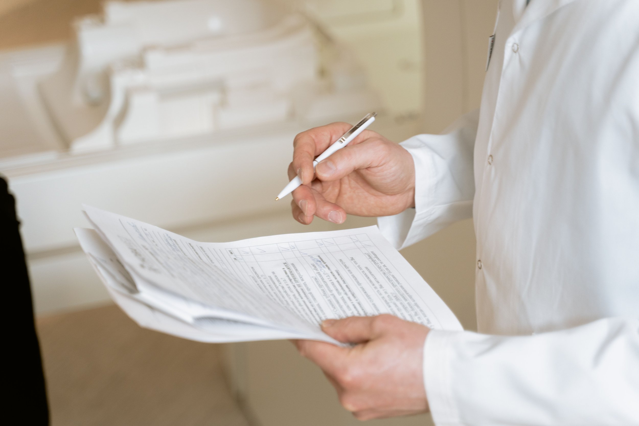 Person in a white shirt holding papers and using a white pen in an office or medical setting with cabinets in the background.