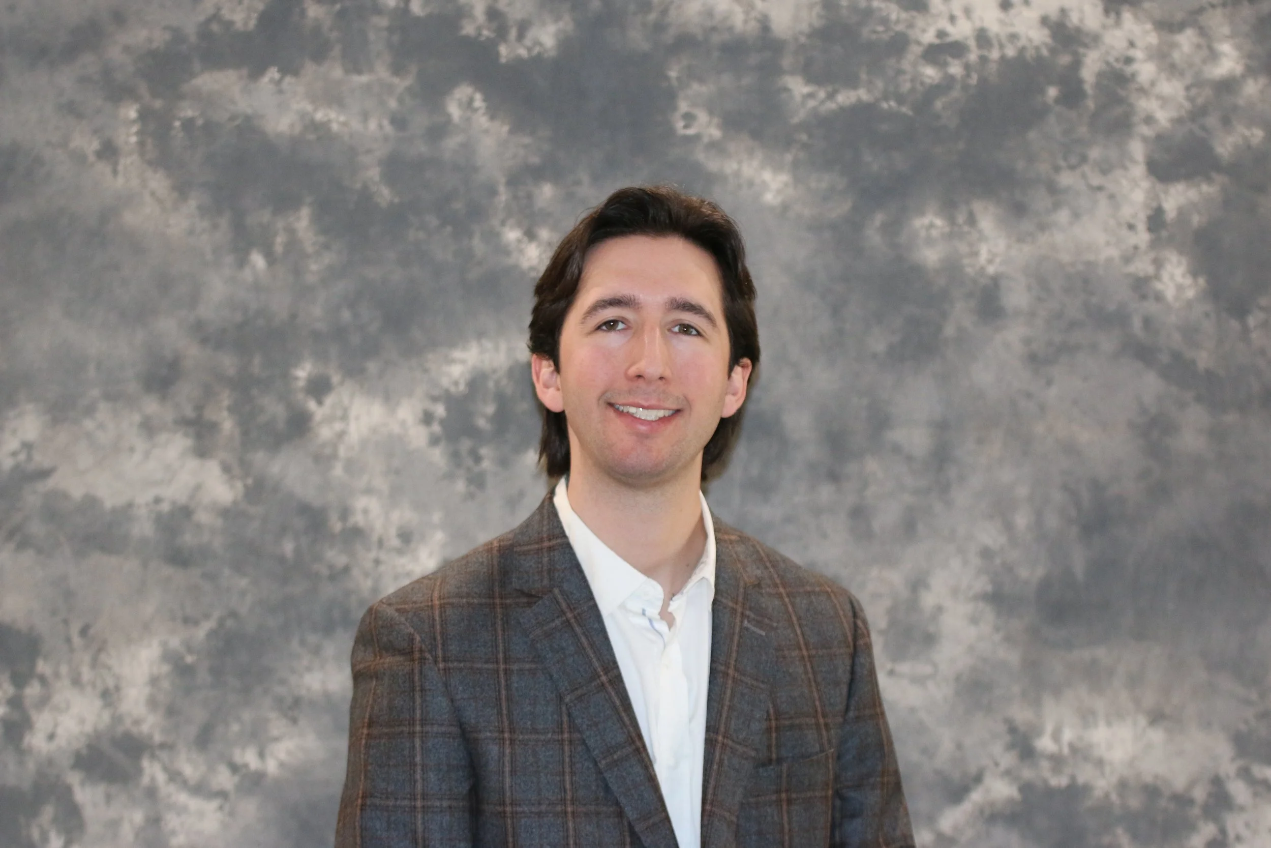 Portrait of a man with dark hair, wearing a brown checkered blazer and white shirt, smiling against a cloudy sky background.
