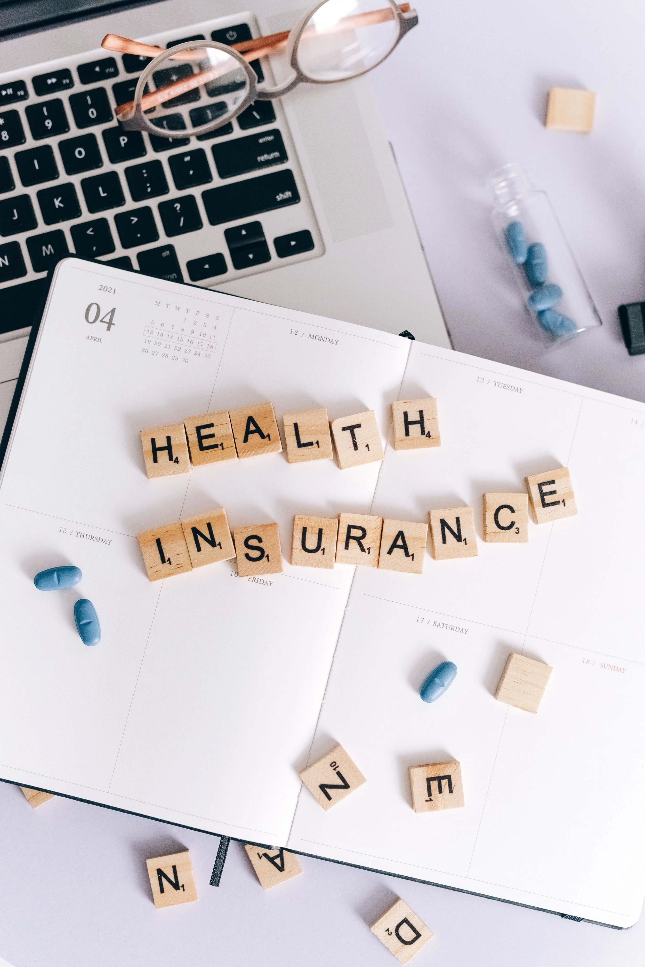 A white planner opened to April 2021 with wooden letter tiles spelling out 'HEALTH INSURANCE' and scattered pills on a white surface. A laptop keyboard, glasses, and a bottle with blue pills are also visible.